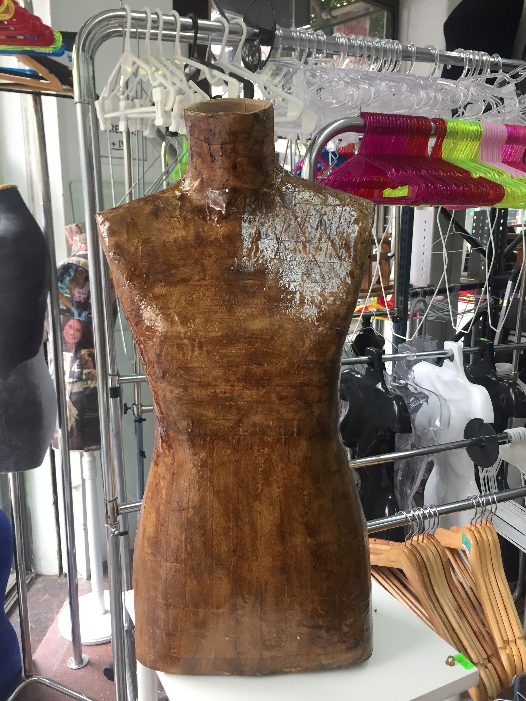 Maniquí de torso marrón de pie en una tienda, rodeado de ropa y estantes.
