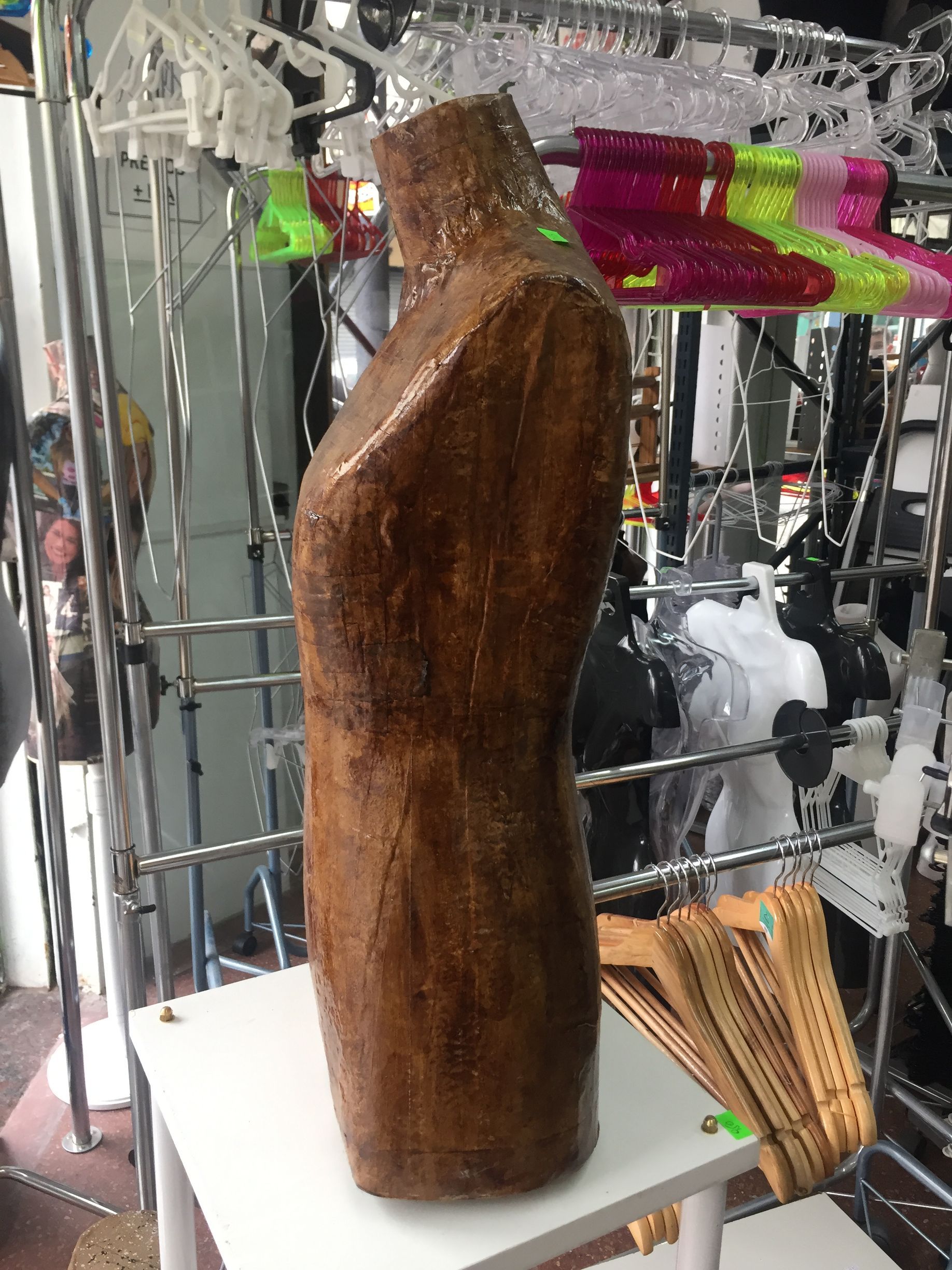 Maniquí de torso marrón sobre una mesa blanca en una tienda, con percheros y perchas de ropa en el fondo.