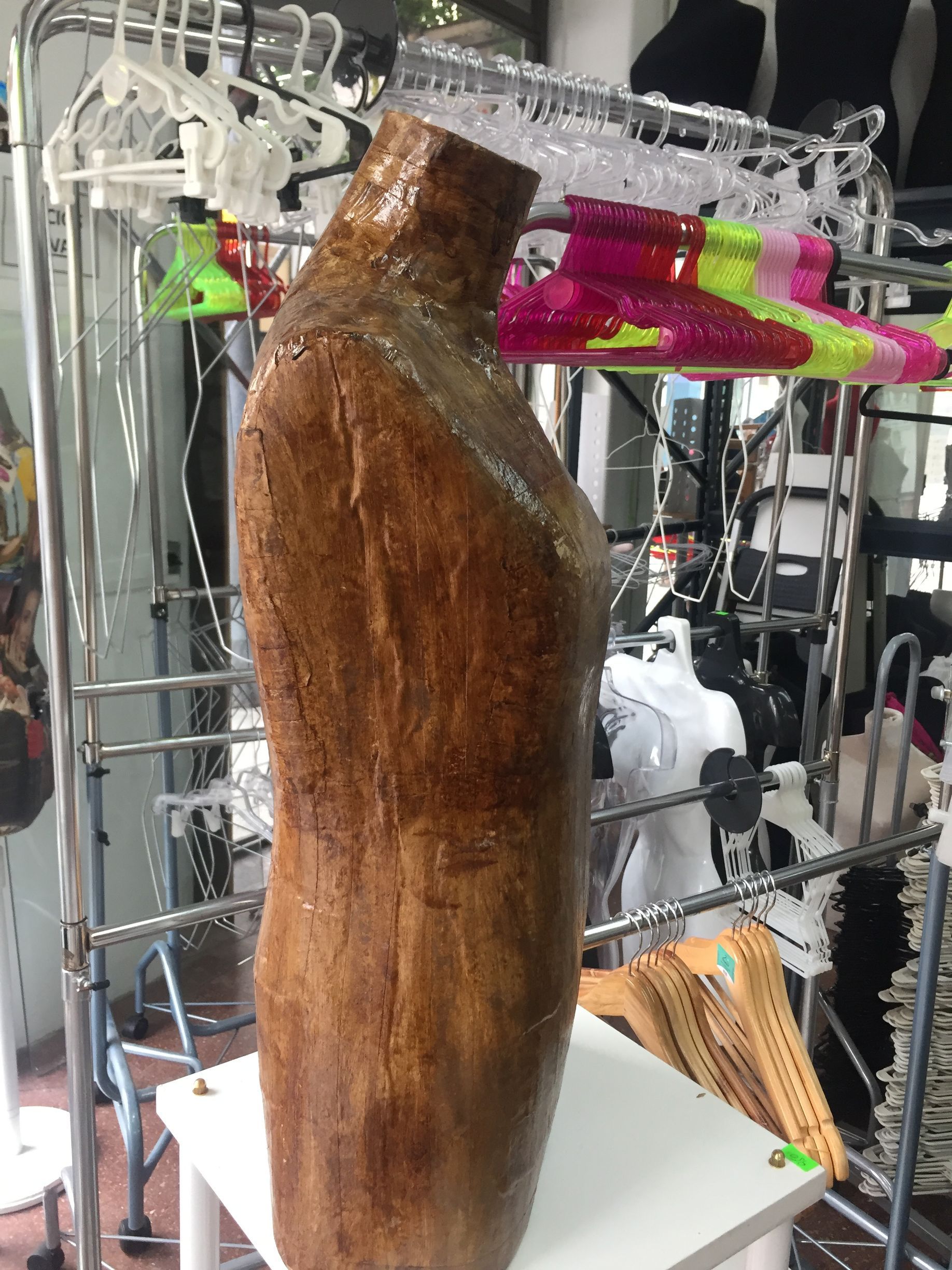 Maniquí de torso de madera de pie sobre una mesa blanca en un entorno minorista con percheros de ropa en el fondo.