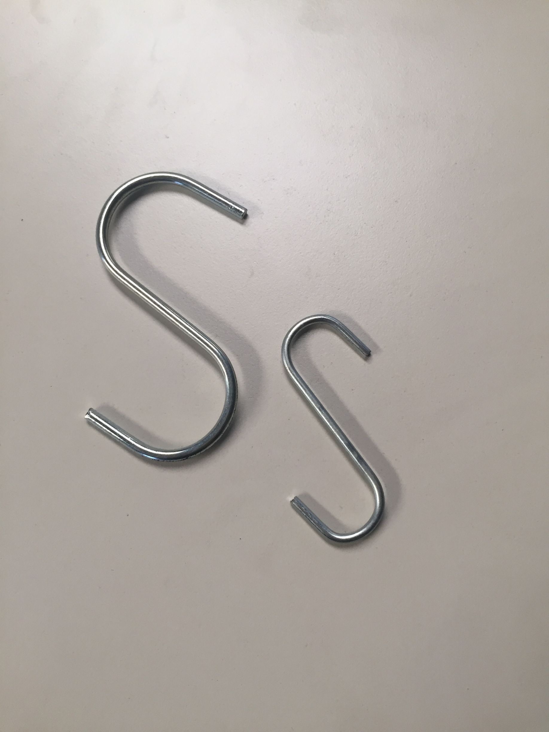 Dos ganchos metálicos en forma de S de diferentes tamaños colocados sobre una superficie clara.