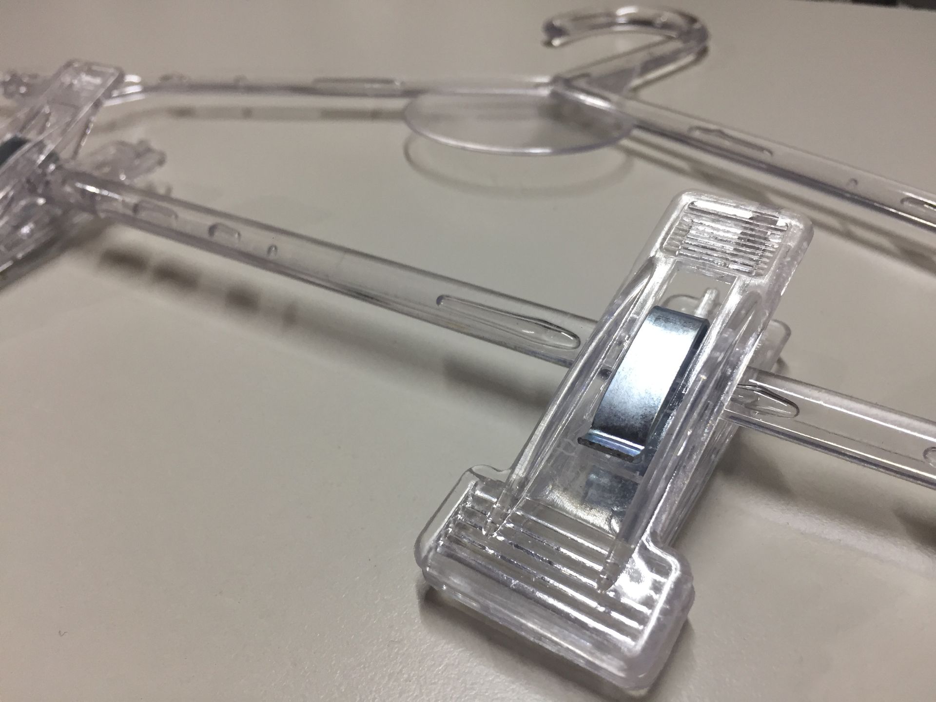 Perchas de plástico transparente para pantalones sobre una superficie blanca, una de ellas enfocada con un clip de metal.