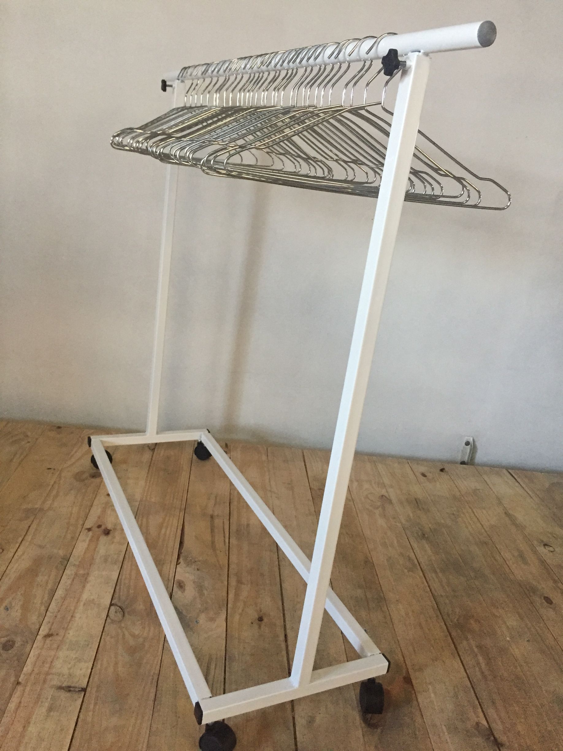 Perchero de metal blanco con ruedas, que contiene numerosas perchas de alambre, sobre suelo de madera.