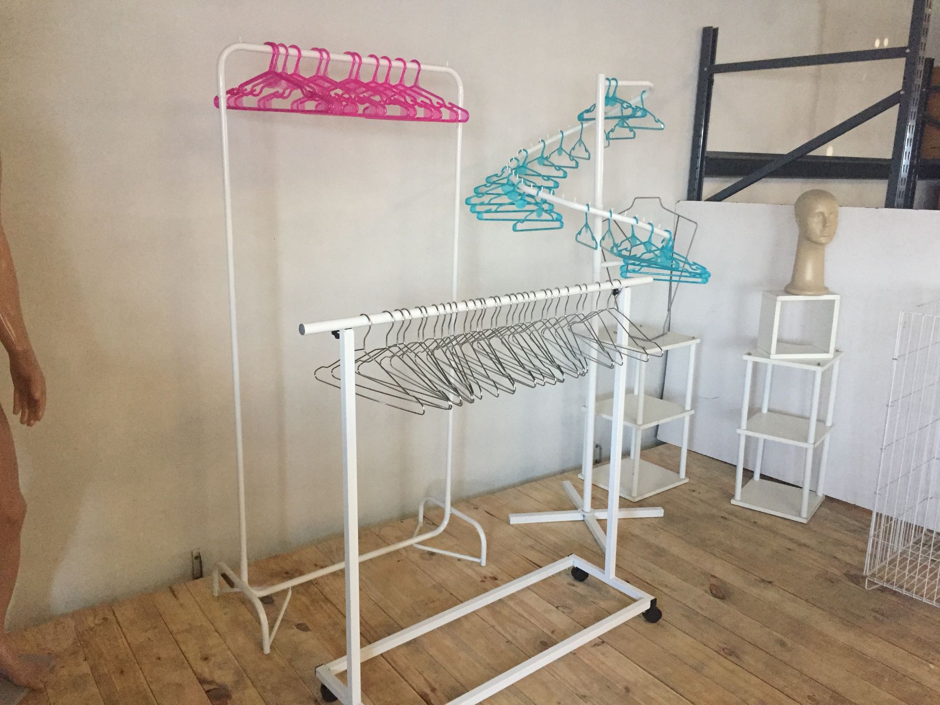 Percheros de ropa de color blanco con perchas de color rosa, verde azulado y negro, colocados contra una pared sencilla.