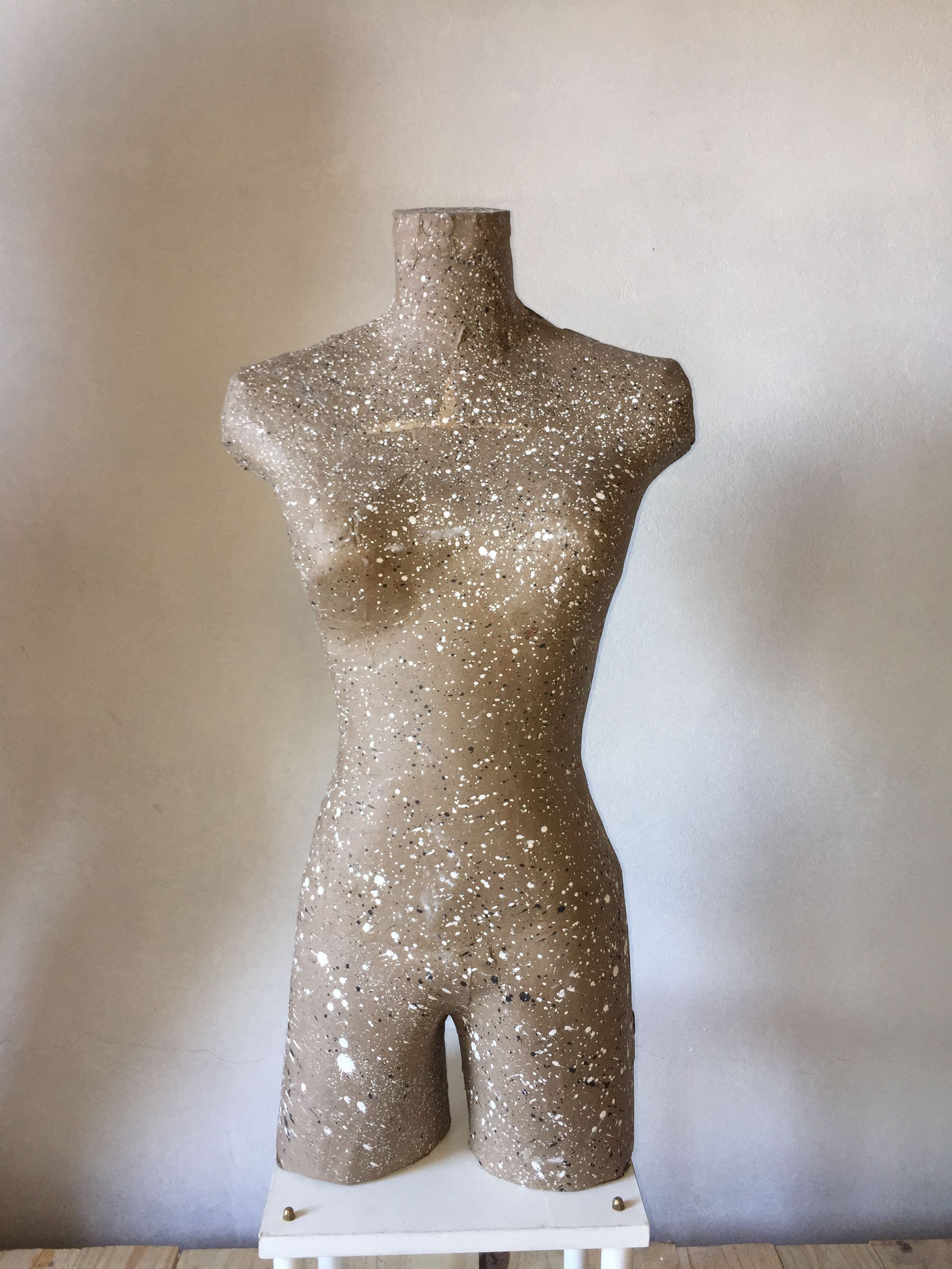 Torso de maniquí cubierto de tela moteada, sobre un soporte blanco. Contra una pared beige claro.