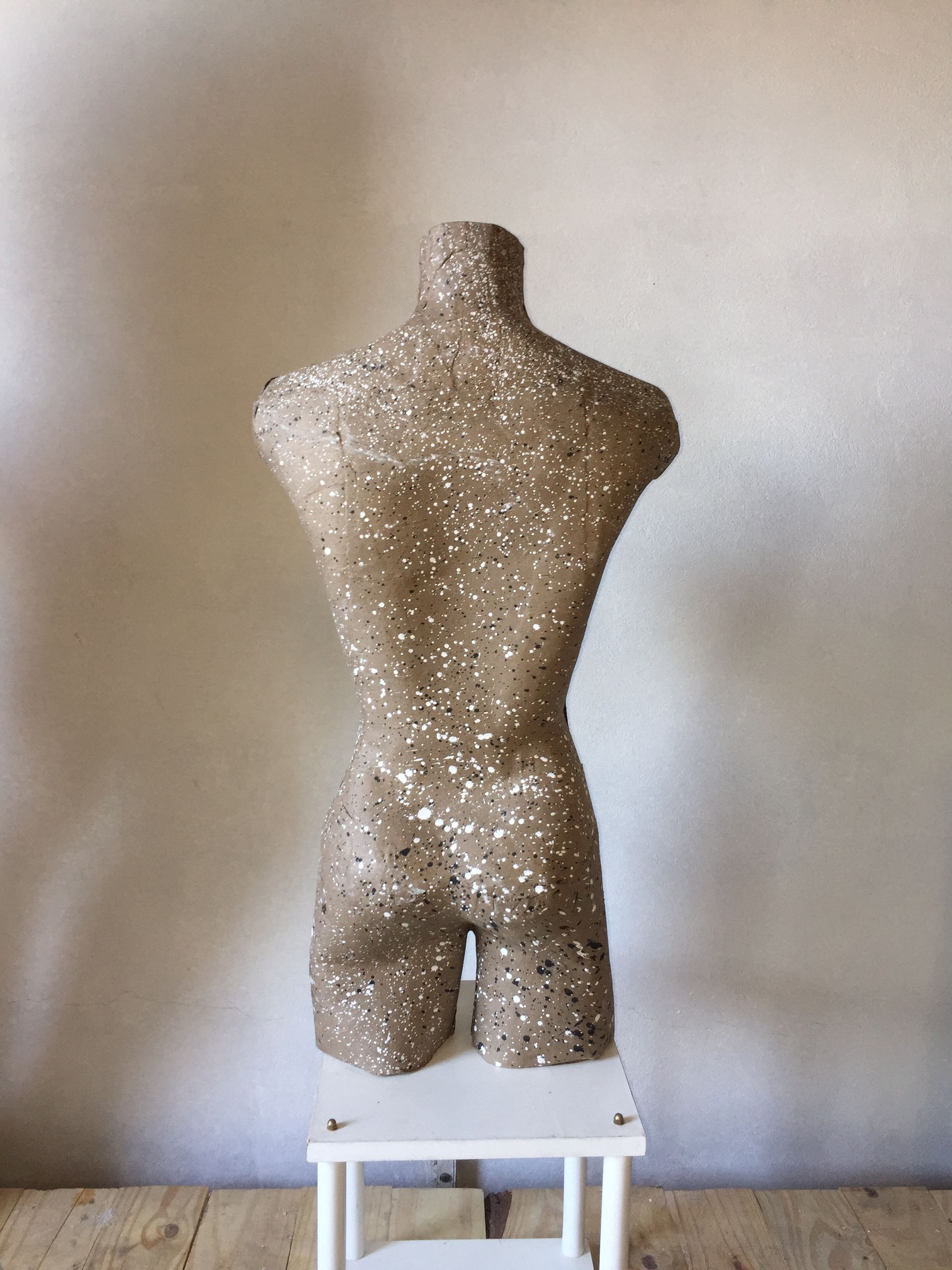 Una escultura de torso con una textura brillante se encuentra sobre un pequeño taburete blanco