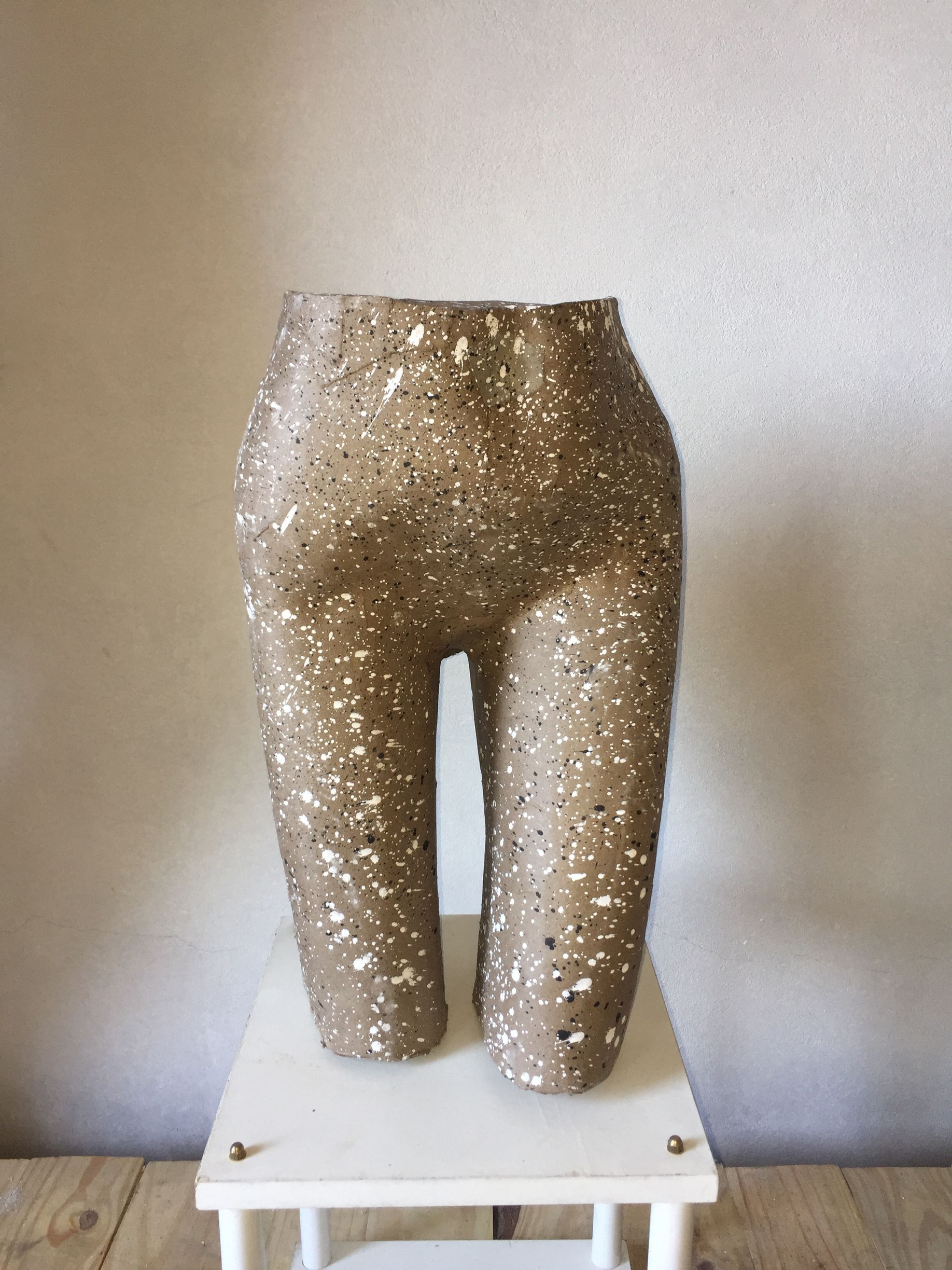 Escultura de piernas y caderas beige sobre una pequeña mesa blanca, contra una pared clara. Textura moteada.