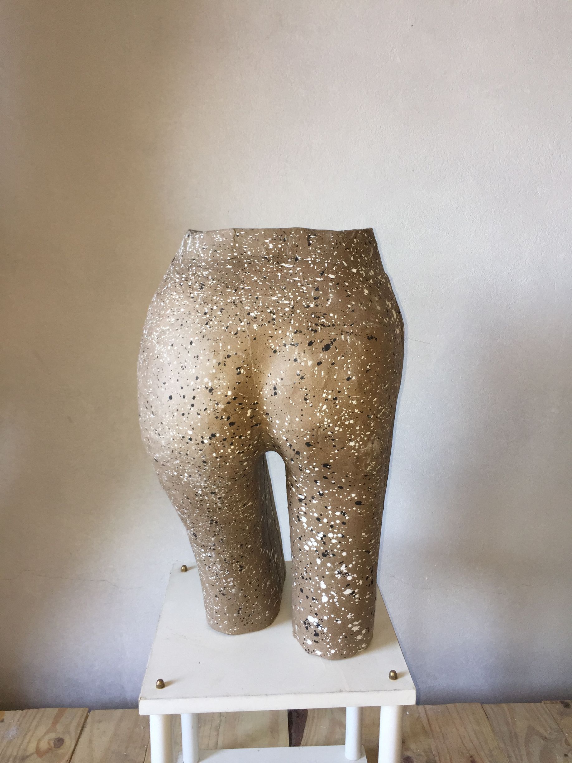 Escultura de piernas y glúteos, recubierta de material moteado en tonos beige y negro, sobre una pequeña mesa blanca.