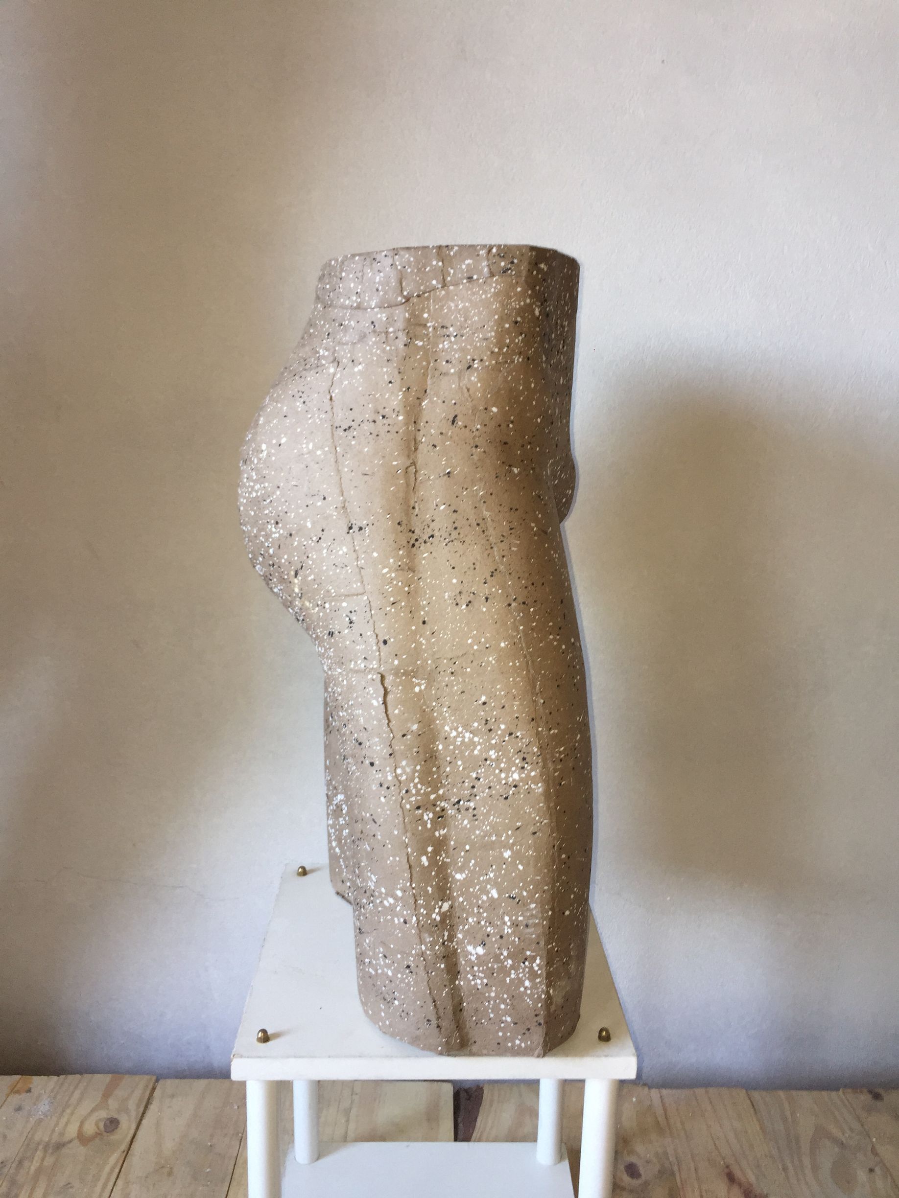 Escultura de una pierna, con textura moteada de color canela, sobre un pequeño taburete blanco, contra una pared clara.