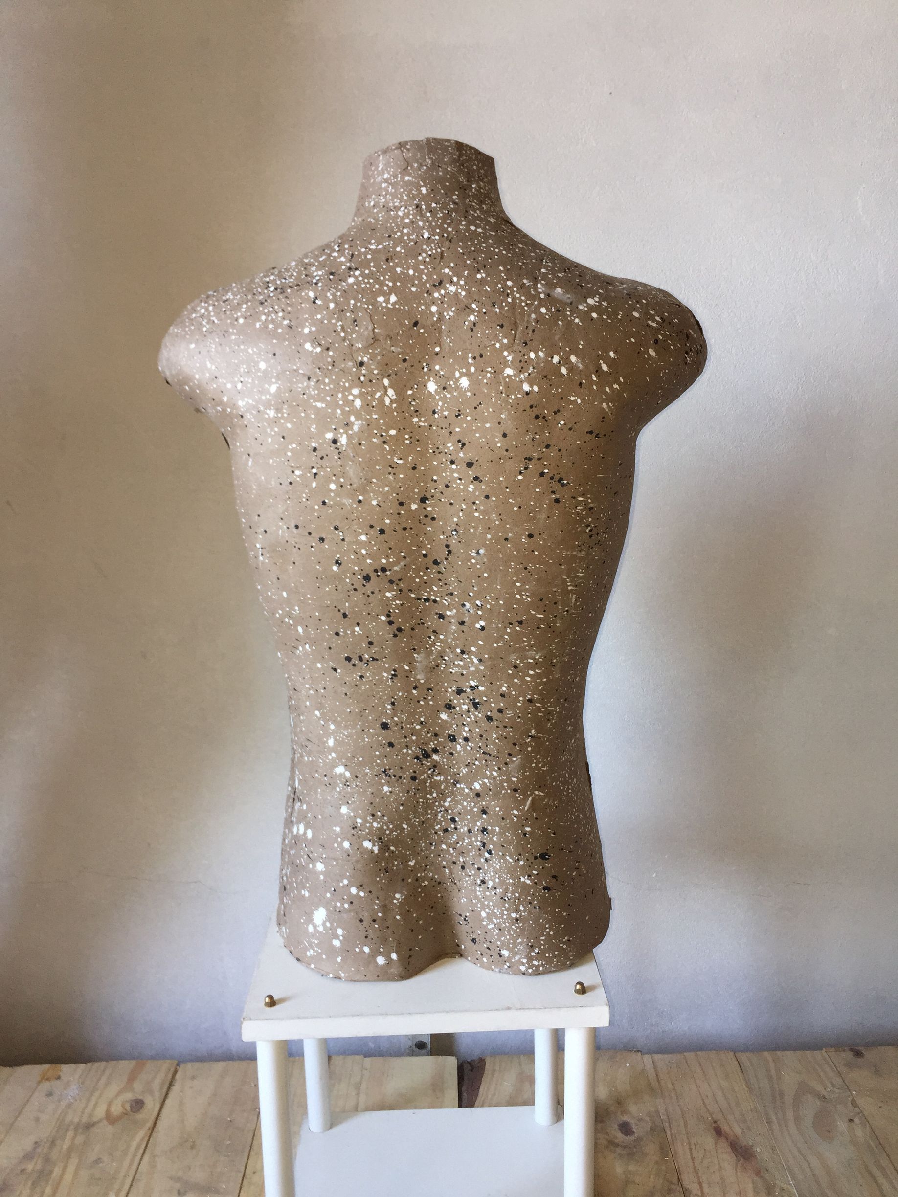 Torso de maniquí con superficie moteada, de pie sobre un pequeño taburete blanco, contra una pared de color crema.