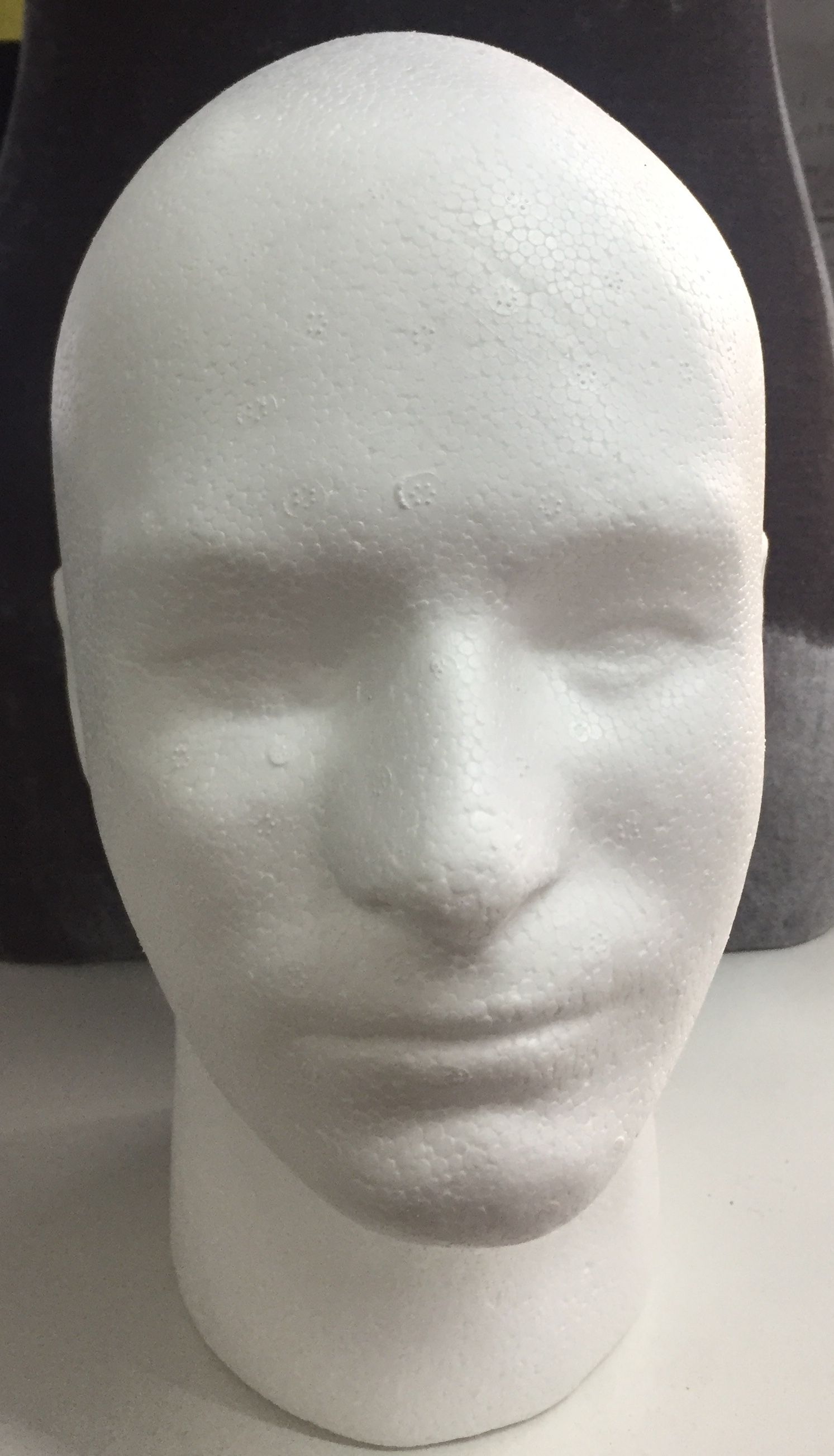 Cabeza de maniquí de espuma blanca, vista frontal. Cabeza ovalada, ojos, nariz y labios cerrados.