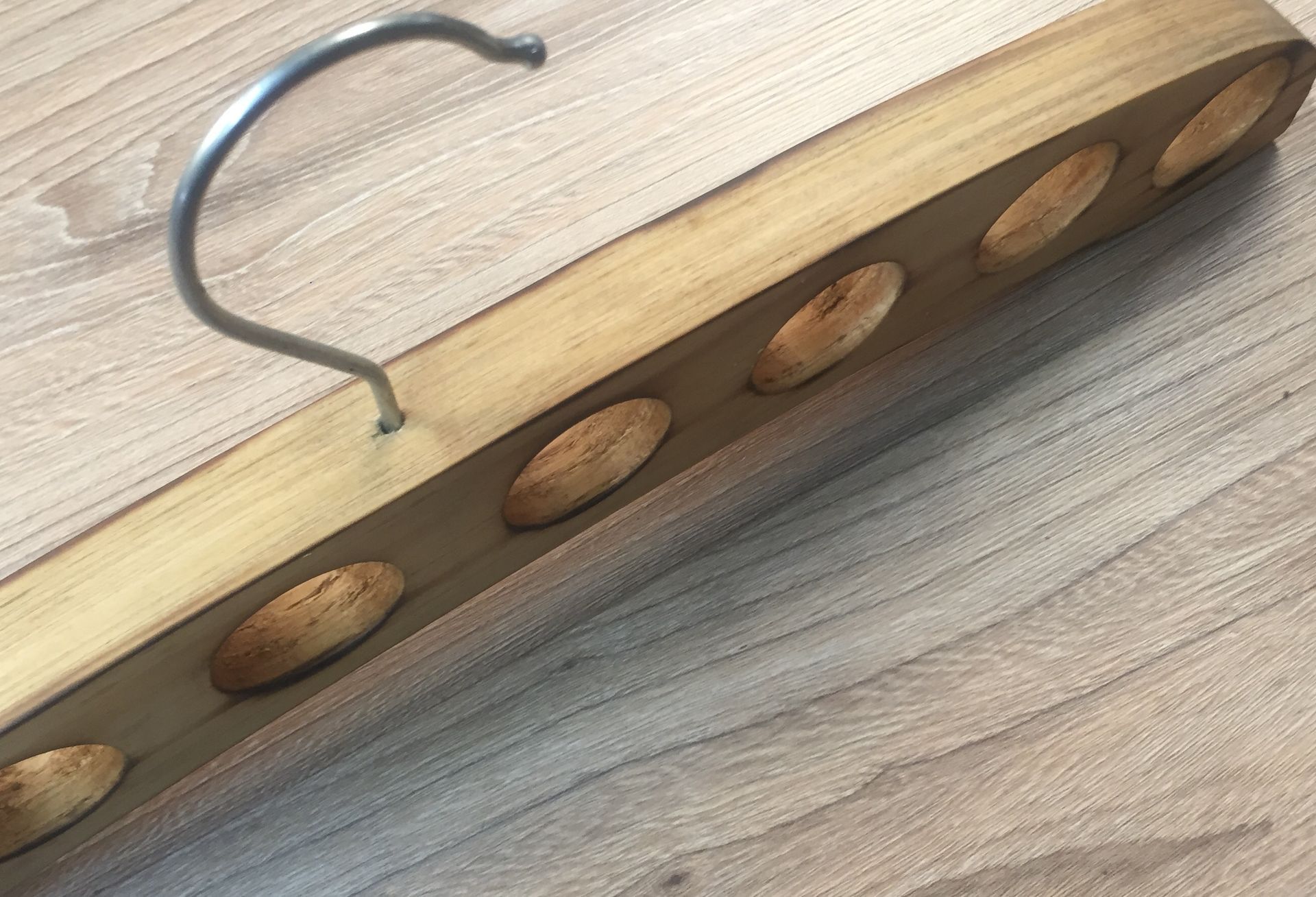Percha de madera con agujeros ovalados y gancho de metal, colocada sobre una superficie de madera.