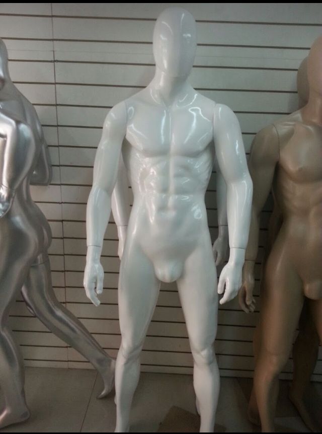 Maniquí masculino de color blanco, acabado brillante, de pie entre otros dos maniquíes frente a un fondo de rejilla.