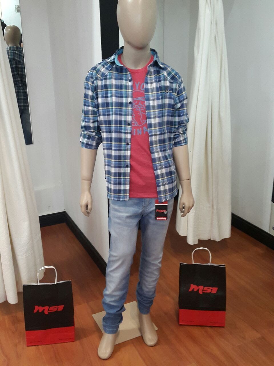Maniquí con una camisa a cuadros sobre una camiseta roja y jeans, en una tienda con bolsas de compras.