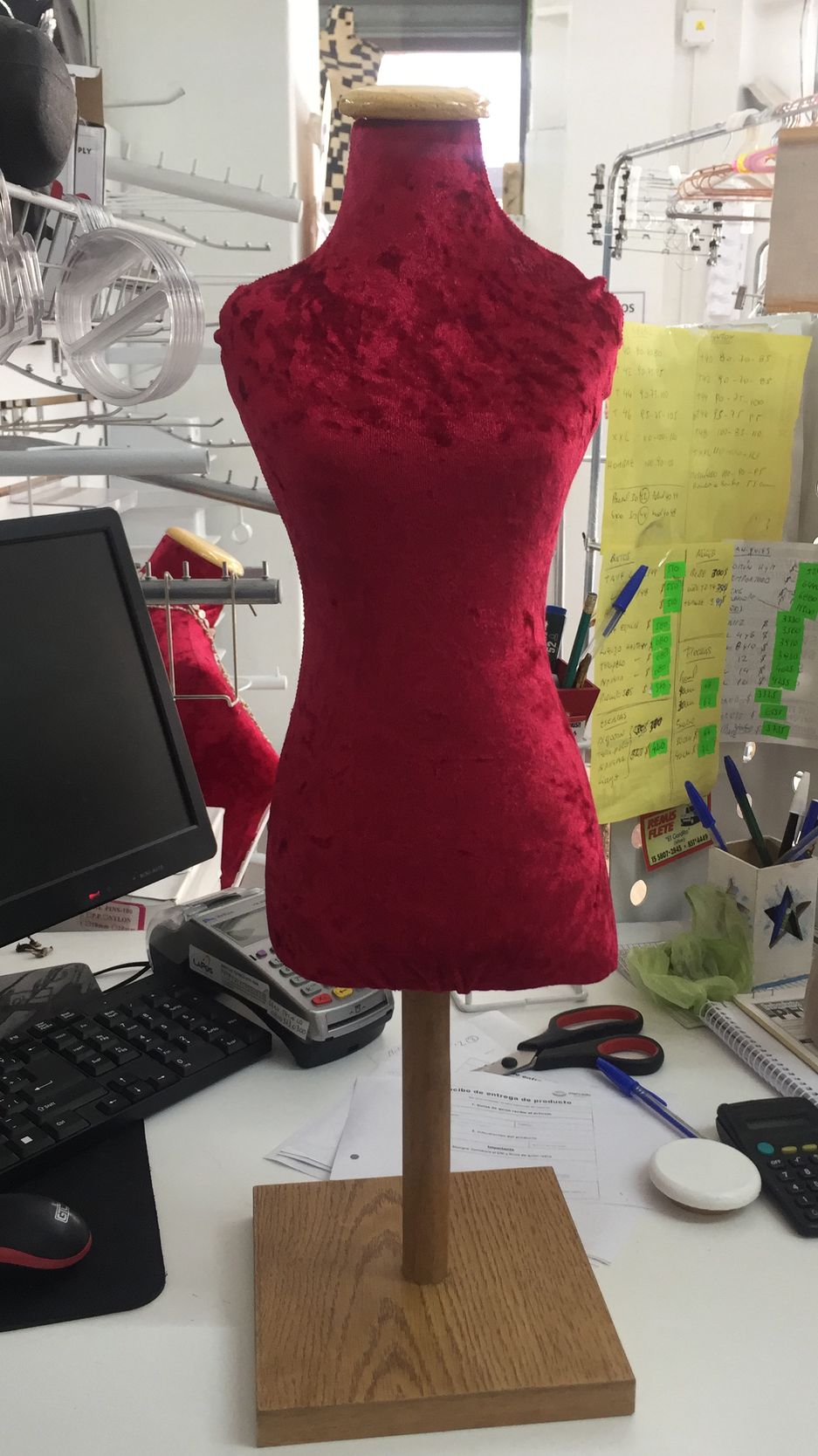 Maniquí de terciopelo rojo sobre soporte de madera, en área de trabajo con computadora y papeles.