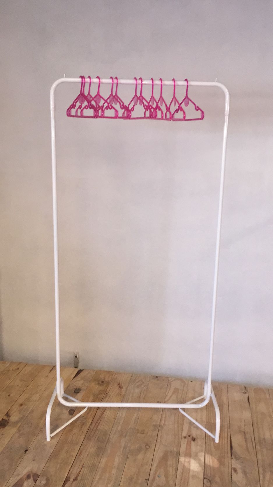 Perchero blanco con perchas rosas, colocado sobre suelo de madera.