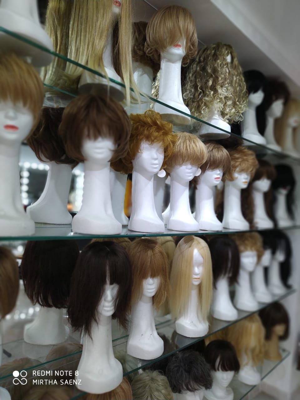 Pelucas sobre cabezas de maniquíes blancos exhibidas en estantes de vidrio dentro de una tienda.