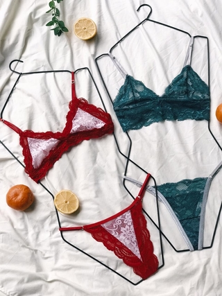 Dos conjuntos de lencería de encaje, rojo y verde, expuestos en perchas de alambre sobre una sábana blanca con frutas.
