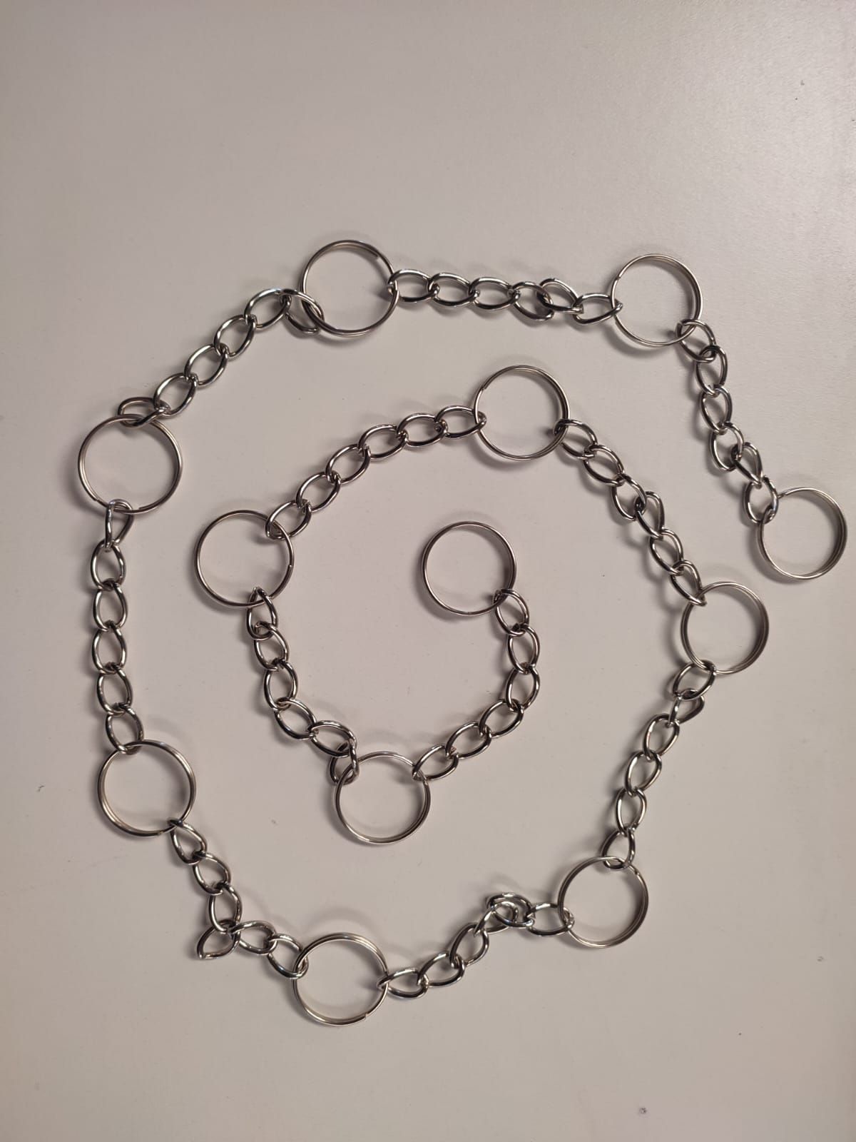 Cadena de metal plateado con anillos conectados dispuestos en patrón de espiral sobre un fondo blanco.