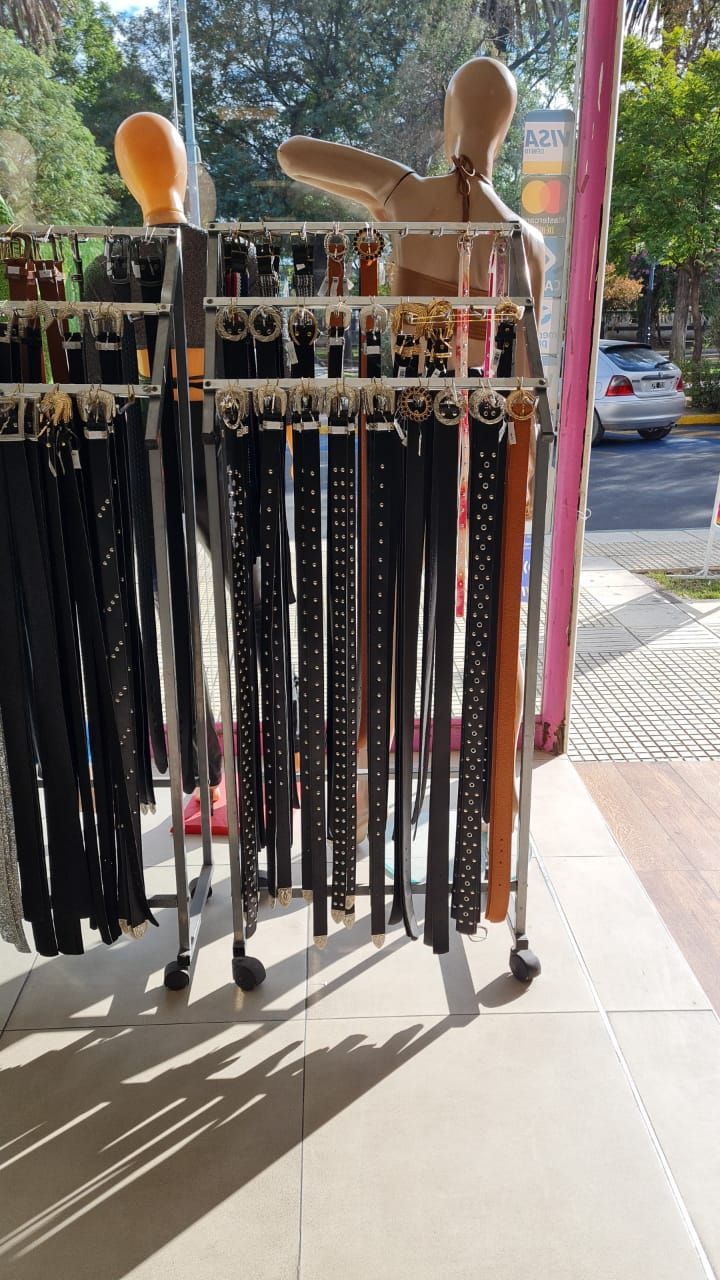 Cinturones expuestos en un perchero con ruedas en una tienda, junto a un maniquí. La luz del sol crea sombras.