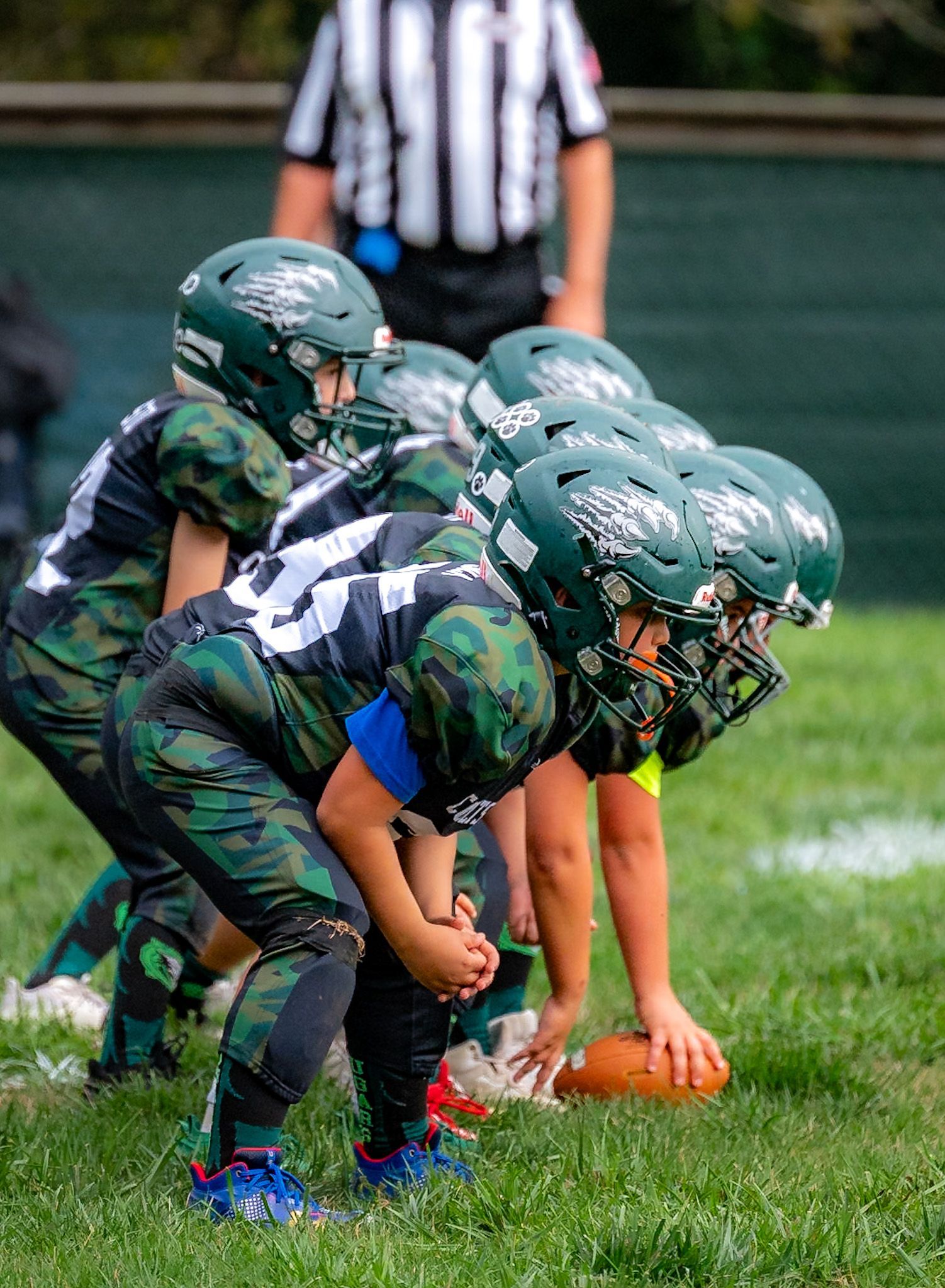 Football players in action on a grassy field; one runs with the ball, pursued by two opponents.