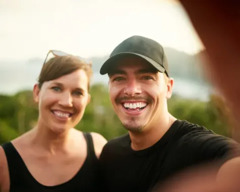 Two smiling people in black shirts and caps taking a selfie outdoors at sunset