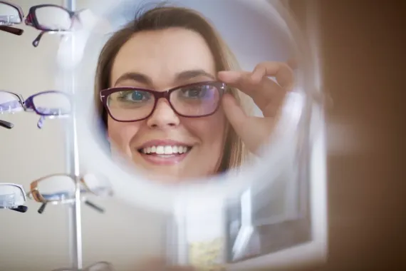Woman adjusting glasses in an eyewear store, smiling at her reflection through a magnifier.