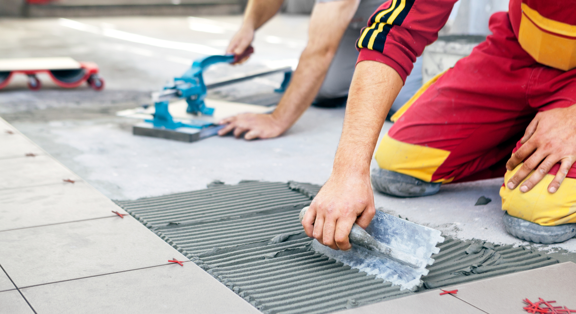 Two people tiling a floor, one spreading mortar with a notched trowel, the other using a tile cutter.