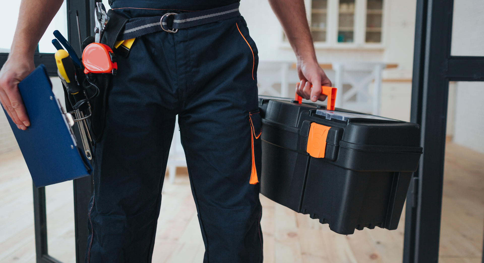 Handyman in navy work pants holding toolbox and clipboard in interior.