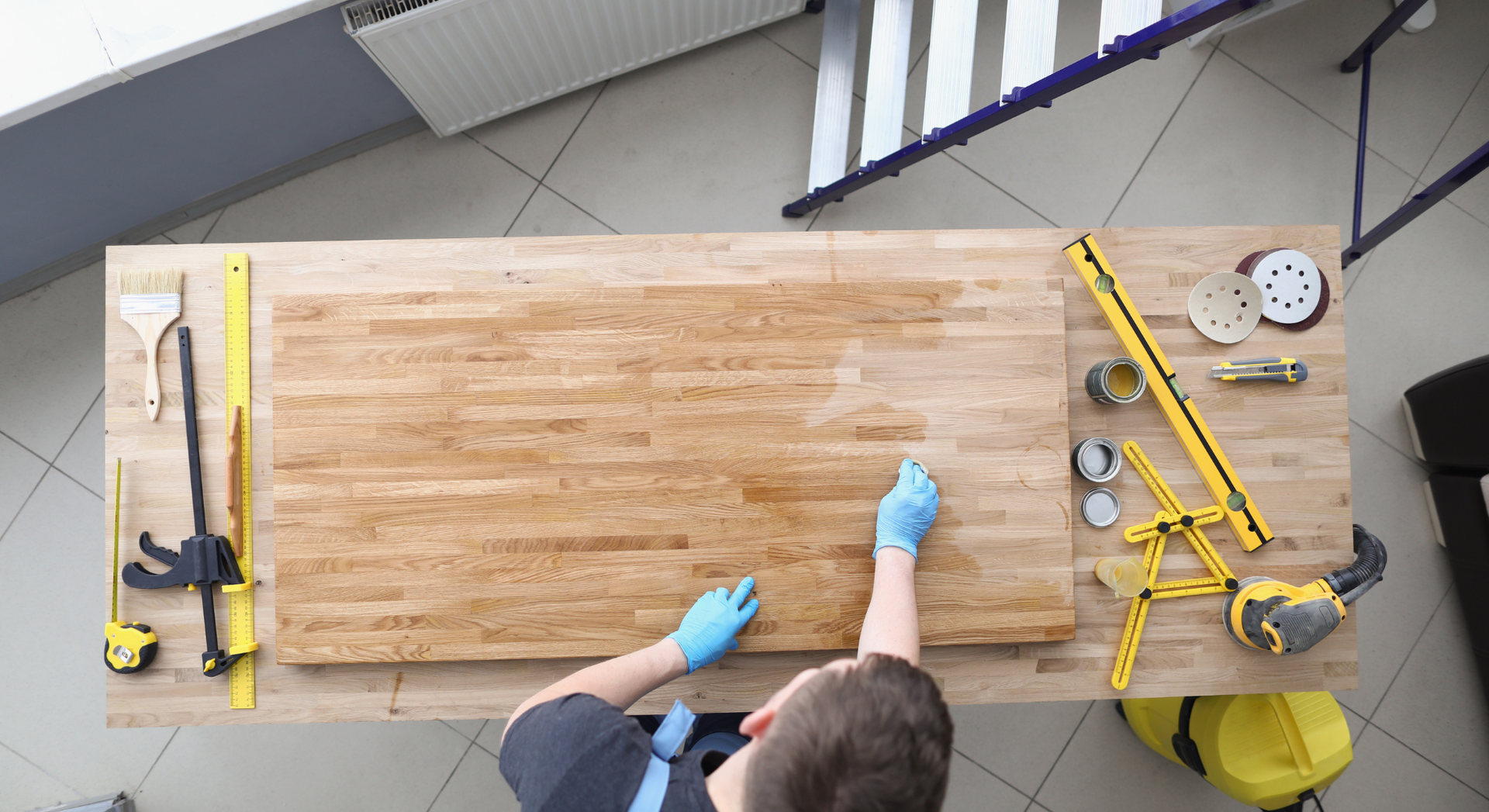 Person applying finish to a wooden tabletop with blue gloves, surrounded by tools on a workbench.