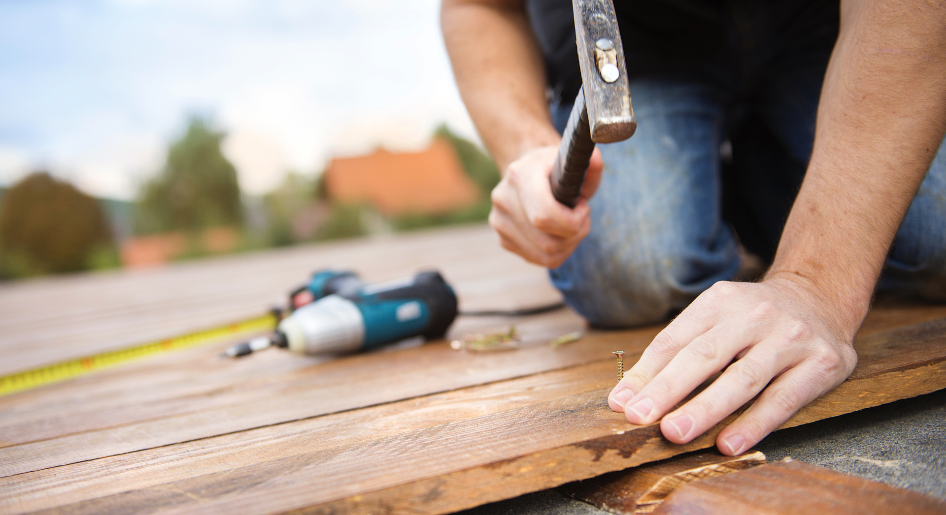 Person hammering a nail into wood on a deck, with a cordless drill and tape measure nearby.