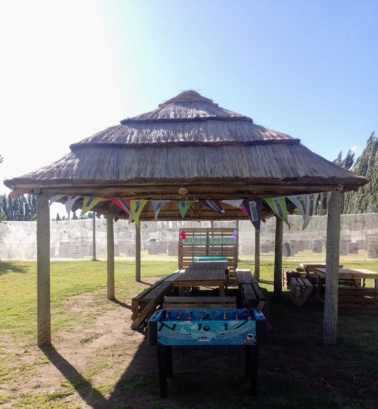 Cenador de madera con techo de paja, mesas de picnic y mesa de futbolín en una zona de césped con banderas.