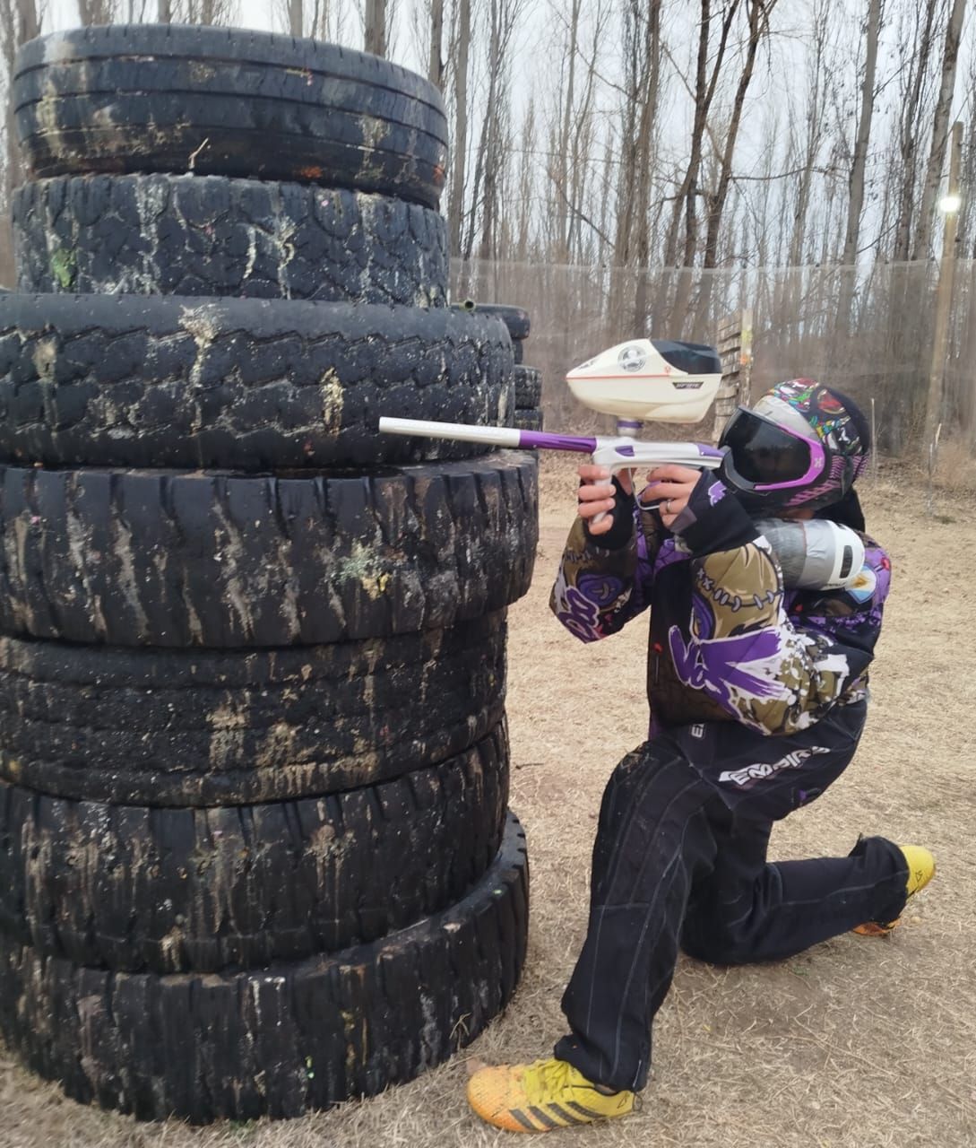 Una persona se arrodilla detrás de una barrera de neumáticos, apuntando con una pistola de paintball violeta y blanca.