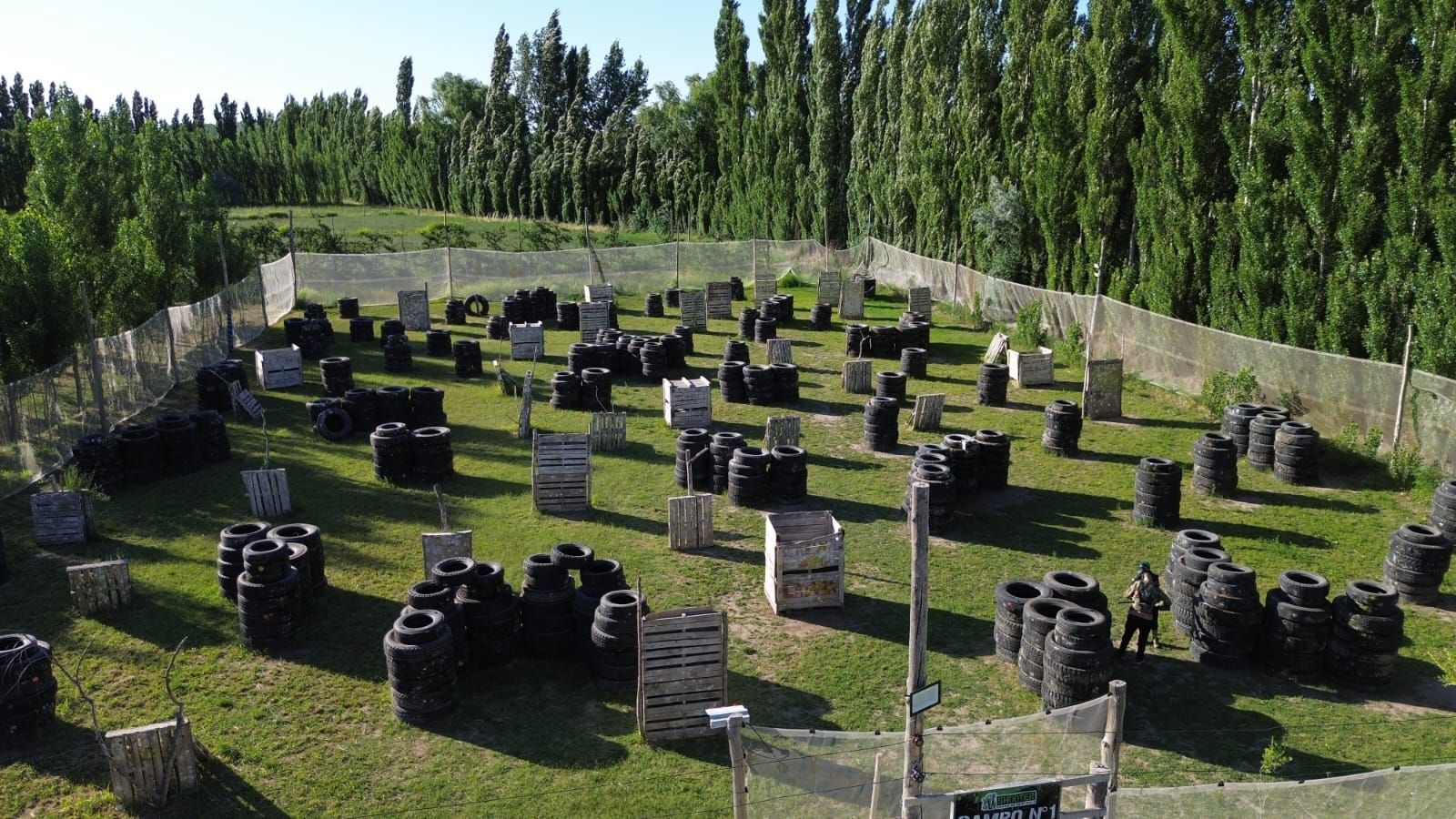 Campo de paintball con obstáculos como neumáticos y estructuras de madera. Césped verde y árboles rodean el área de juego.