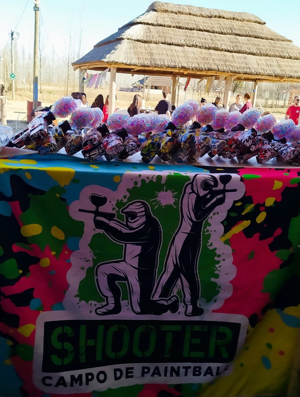 Trofeos de paintball y logotipo sobre una mesa, jugadores y estructura de techo de paja al fondo.