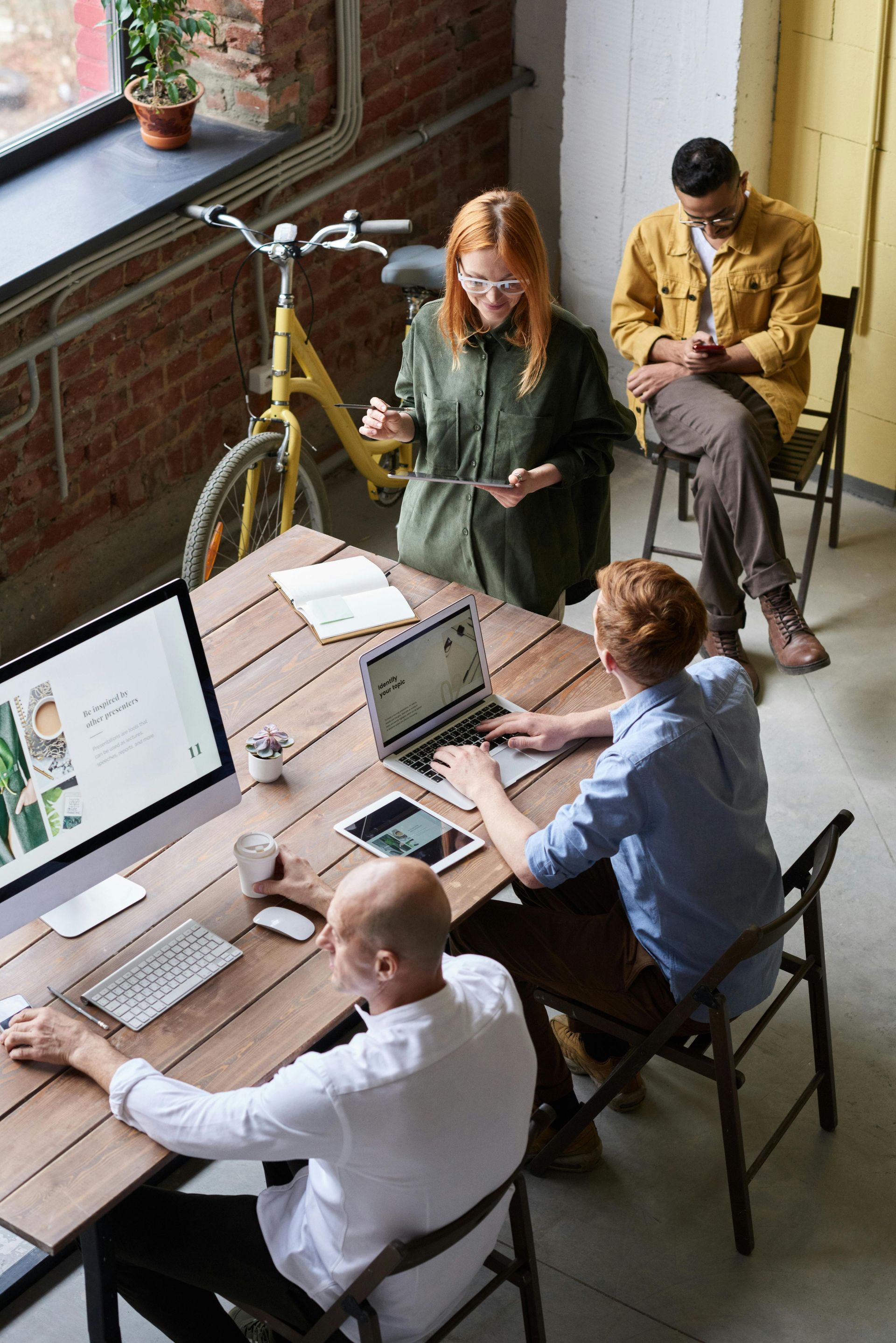 People working at a table, some using computers, others looking on. Office setting with a bicycle.