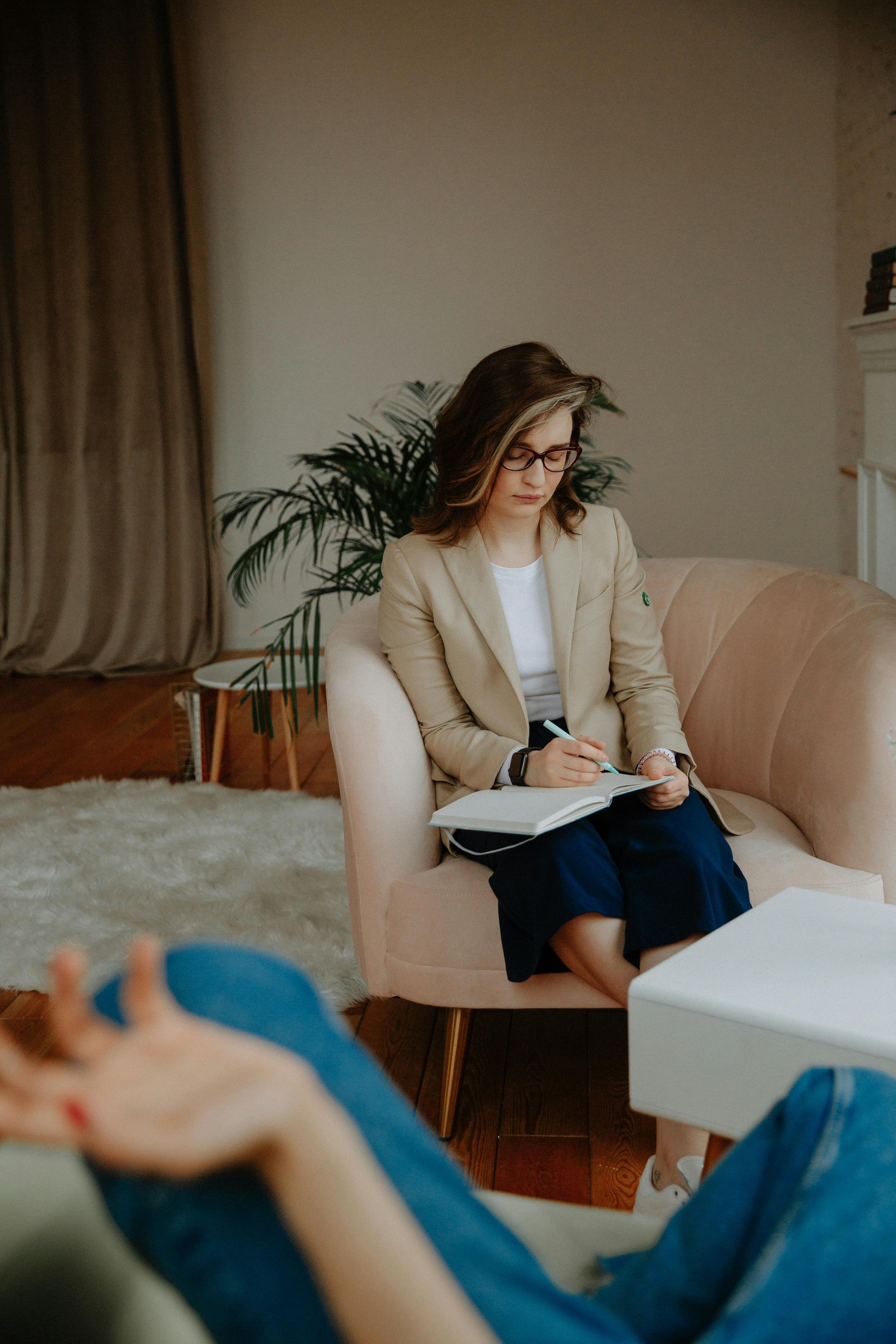 A therapist in a beige blazer takes notes while the patient reclines, arm raised, during a session.