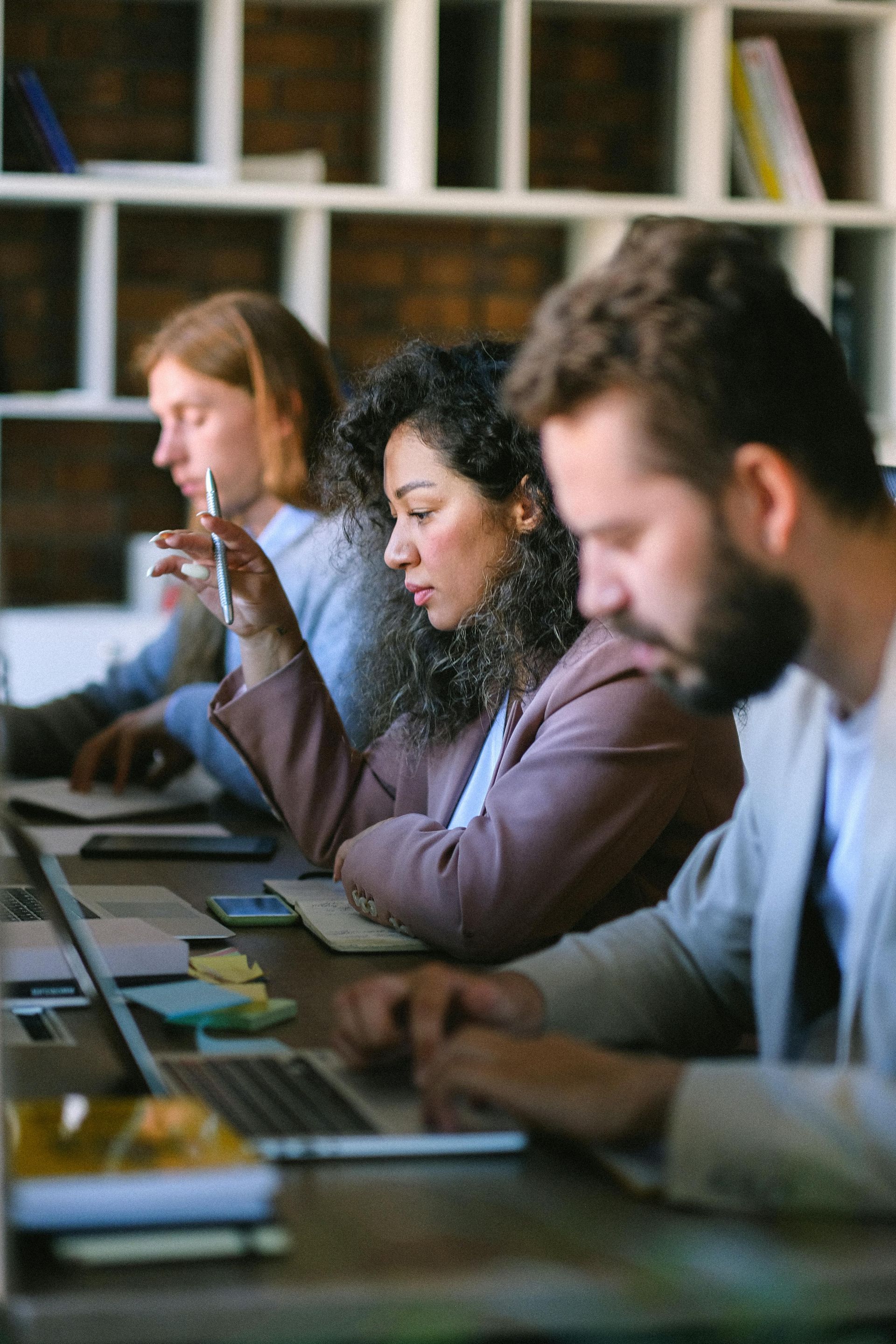 Three people working at a long table with laptops in an office.