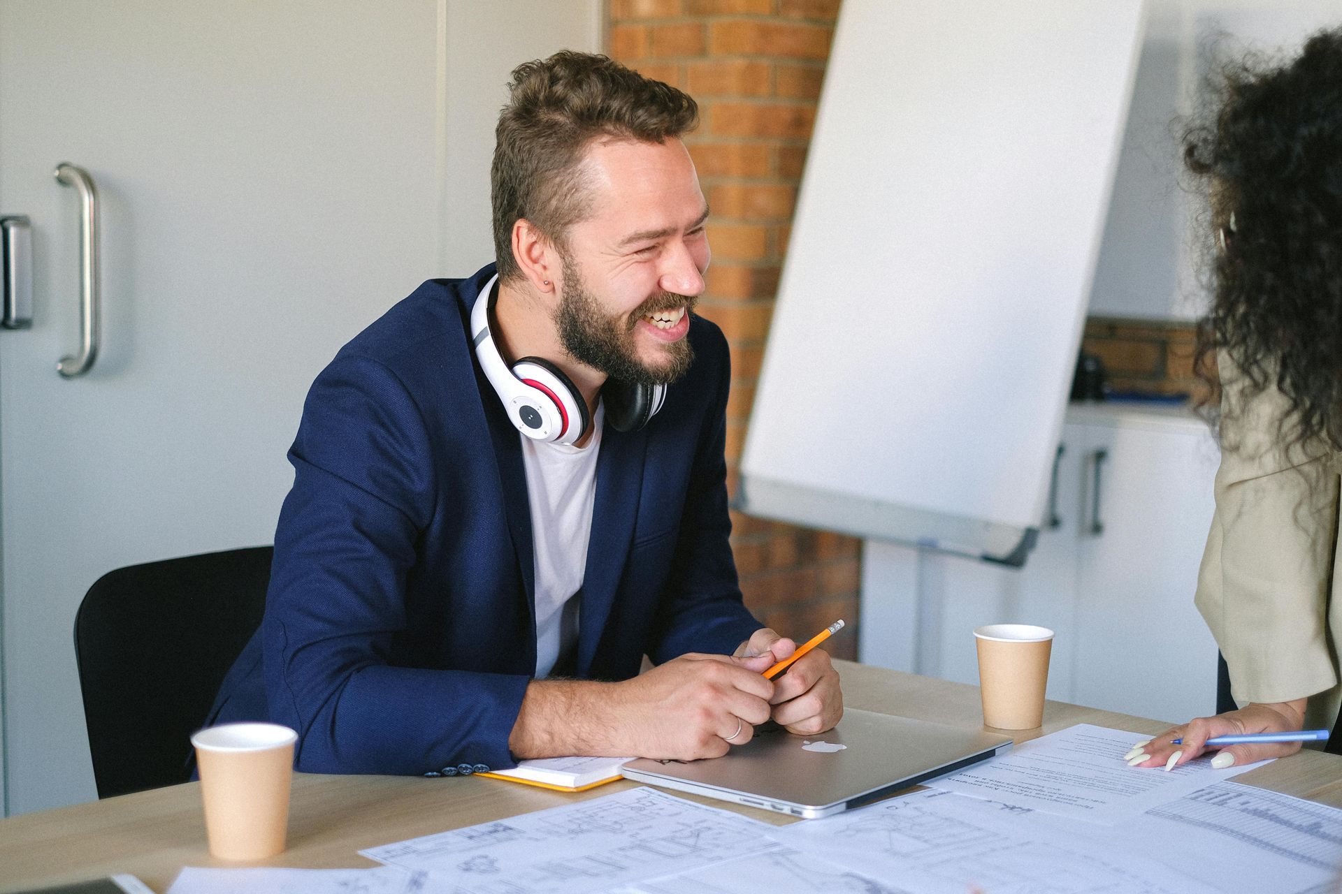 Man in a blue blazer smiles during a meeting, with headphones, paper cups, and a laptop on the table.