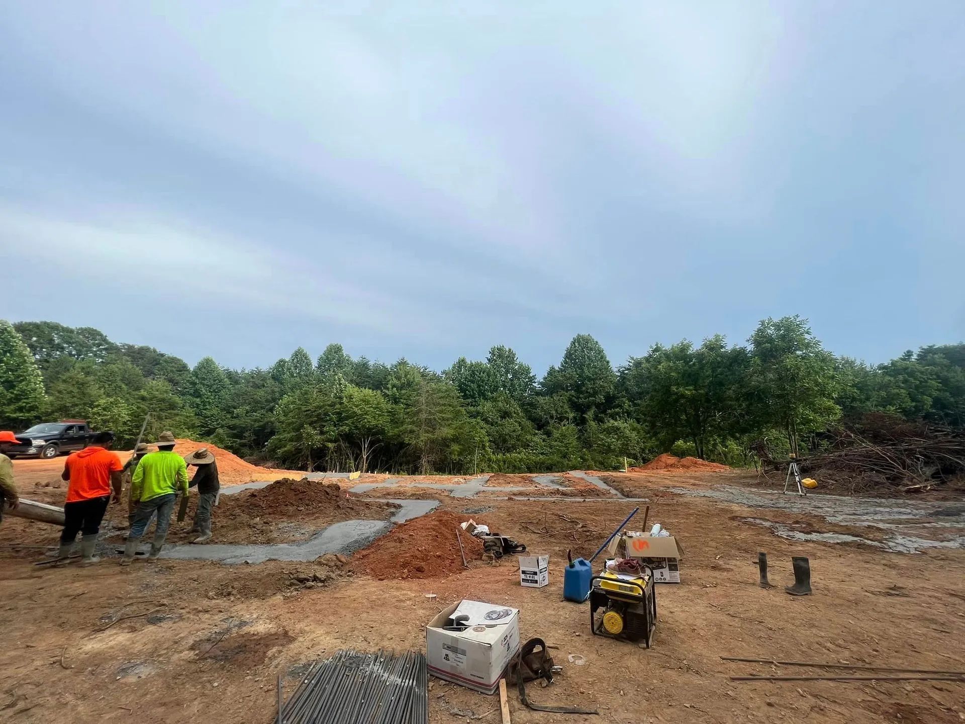 Construction site with workers, foundation outline, tools, and trees under a cloudy sky.