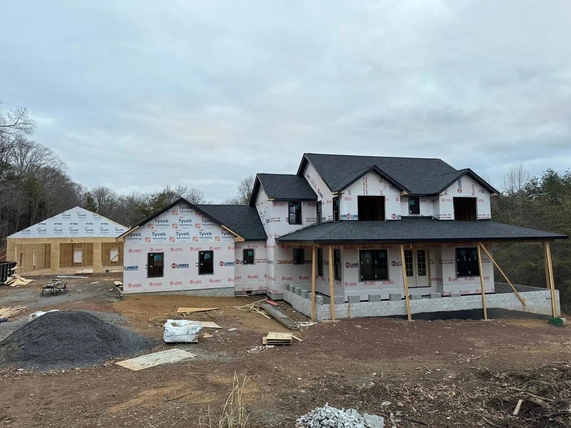 House under construction with detached garage, covered in protective wrap and dark roof, cloudy day.