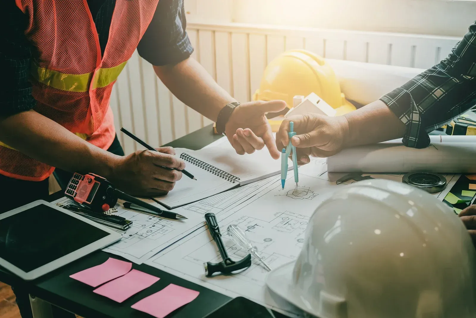 Two people reviewing blueprints at a desk with tools, hard hats, and a tablet, indoors.