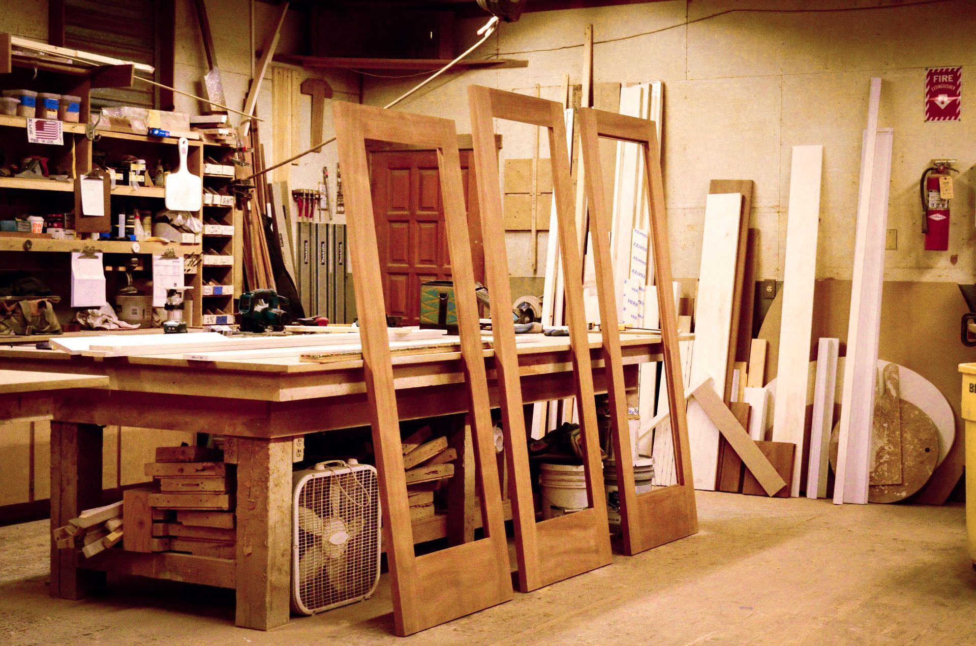 Woodworking shop with four door frames leaning on a workbench and stacks of wood.