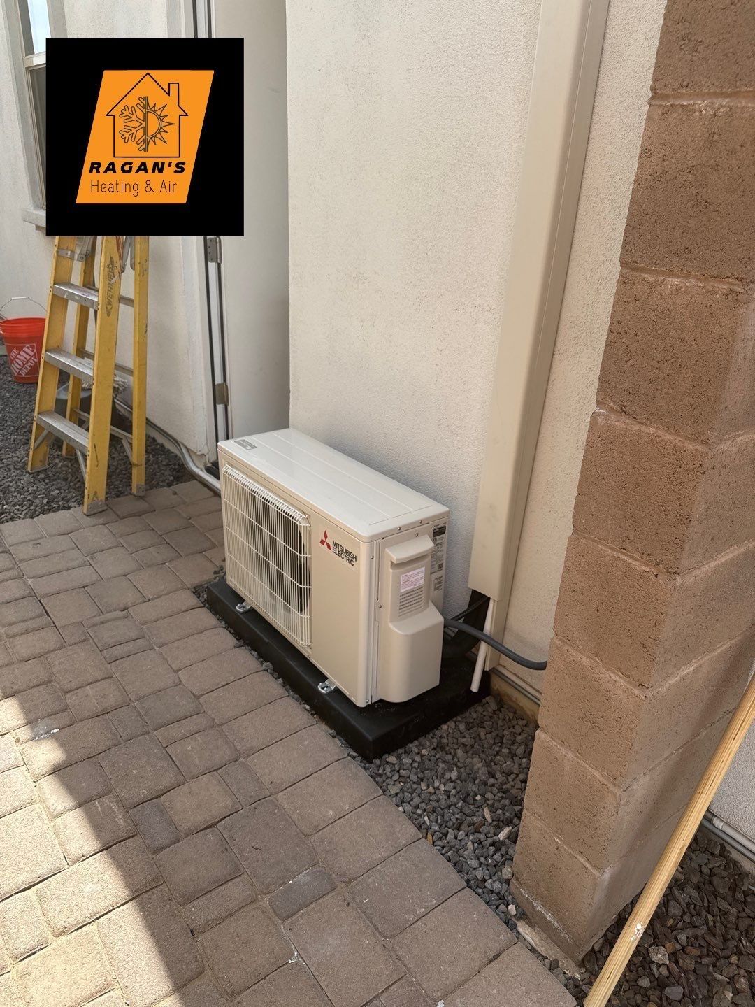 An outdoor air conditioning unit on a black platform next to a white wall and a brick column.