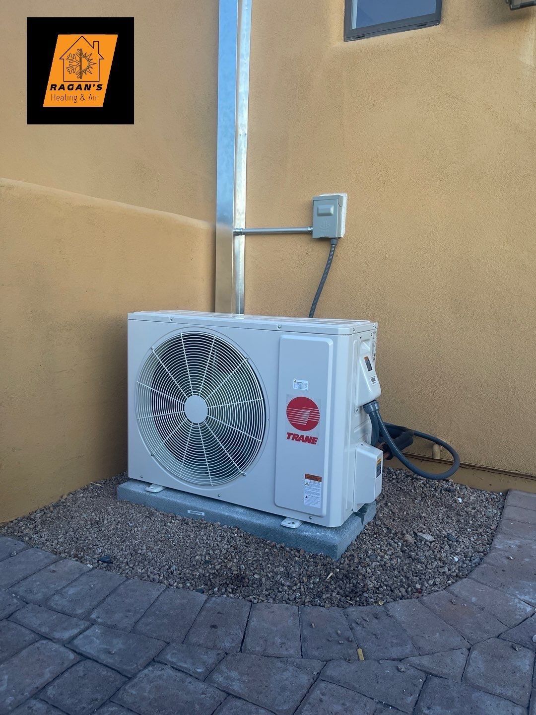Outdoor air conditioning unit against a light brown wall, with a small gravel base on a brick patio.