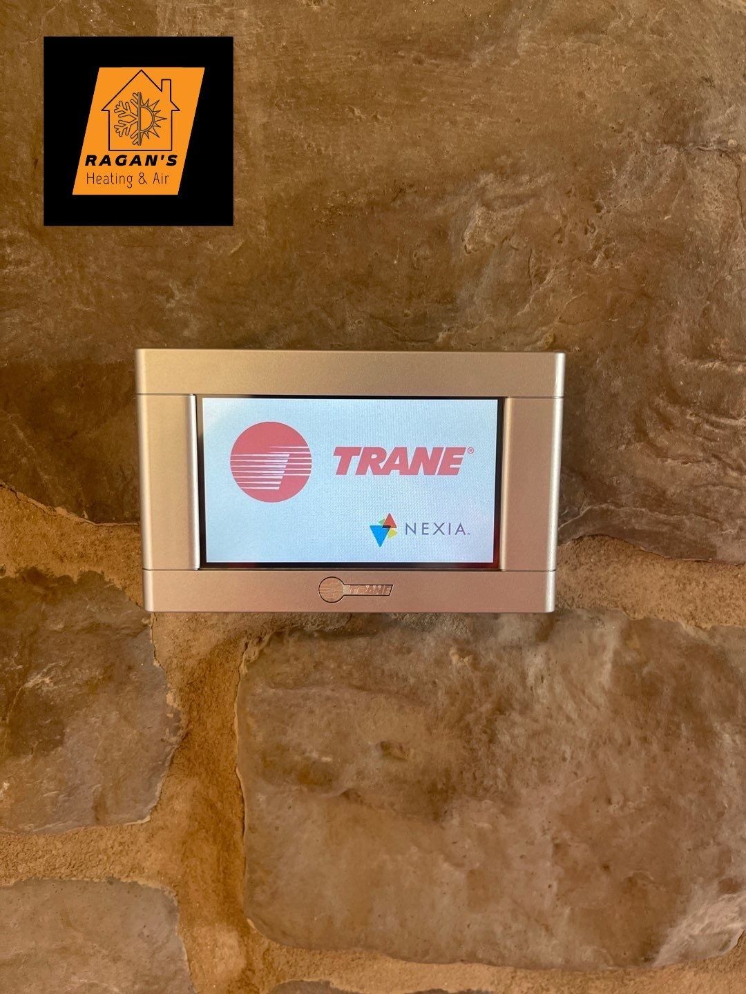 Trane thermostat display on a stone wall. The Trane logo is visible. A company logo is above the thermostat.