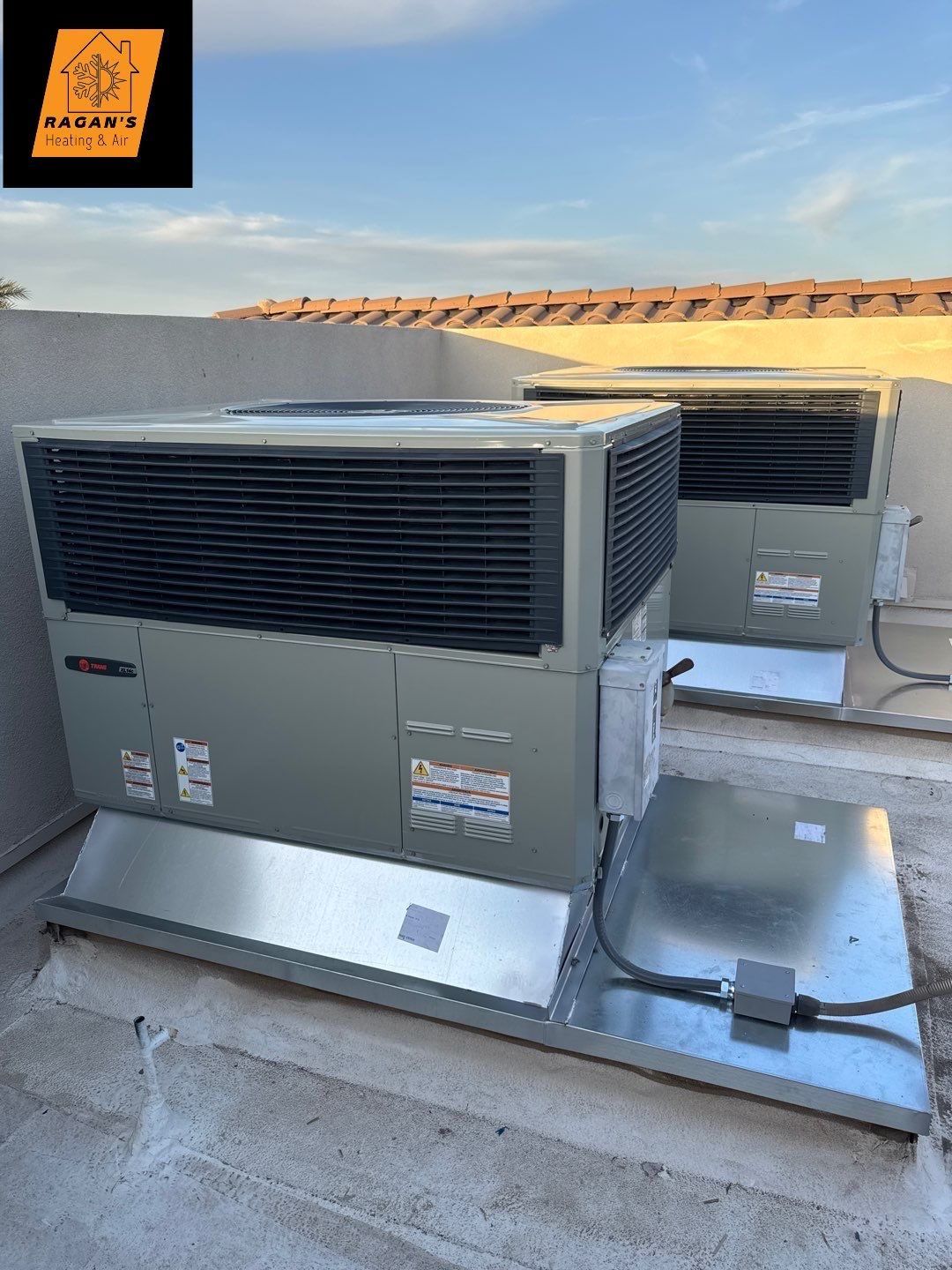 Two rooftop HVAC units, gray with black vents, sit on metal platforms. Blue sky background.