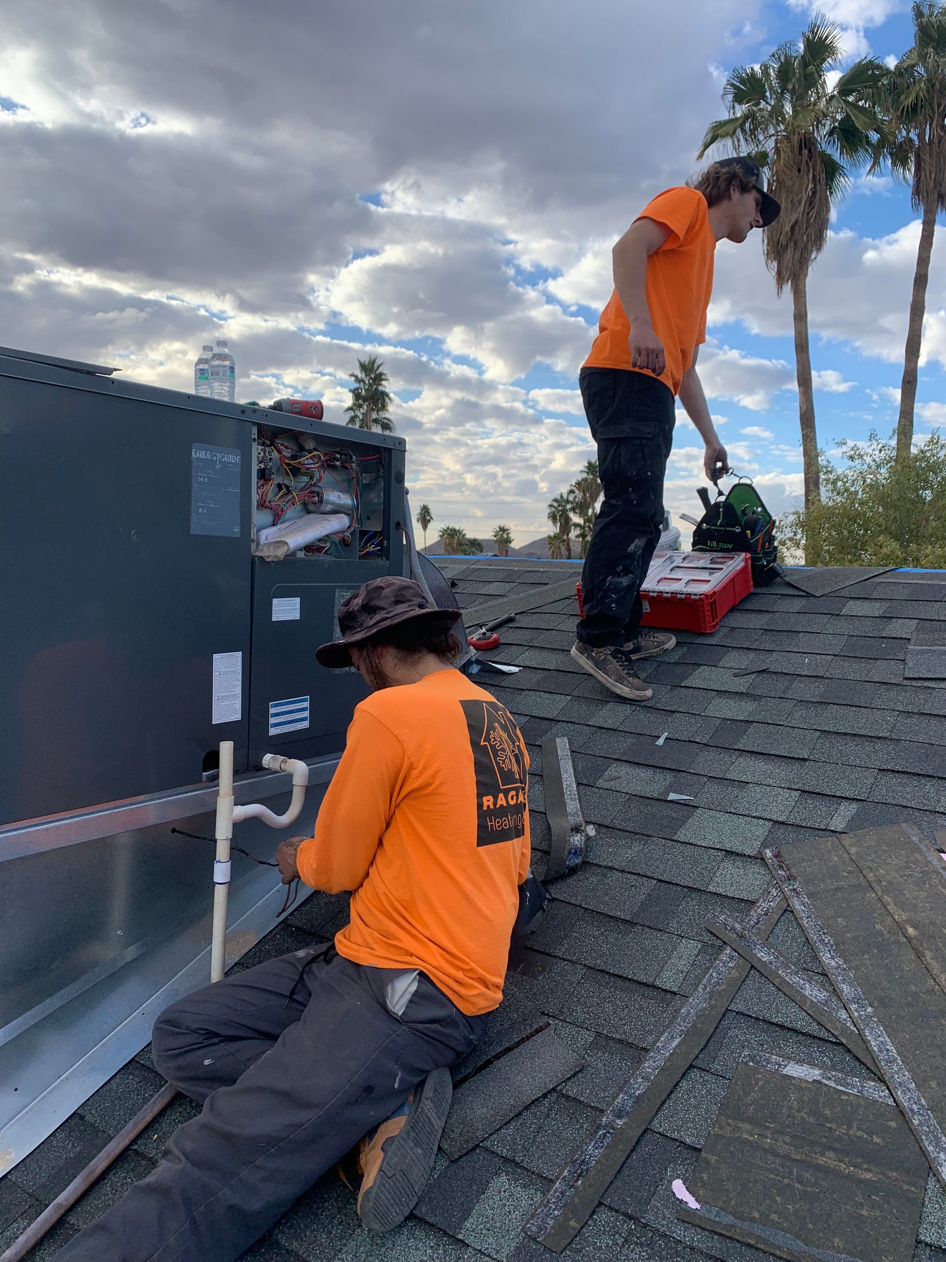 Two HVAC technicians in orange shirts work on rooftop equipment under a cloudy sky.