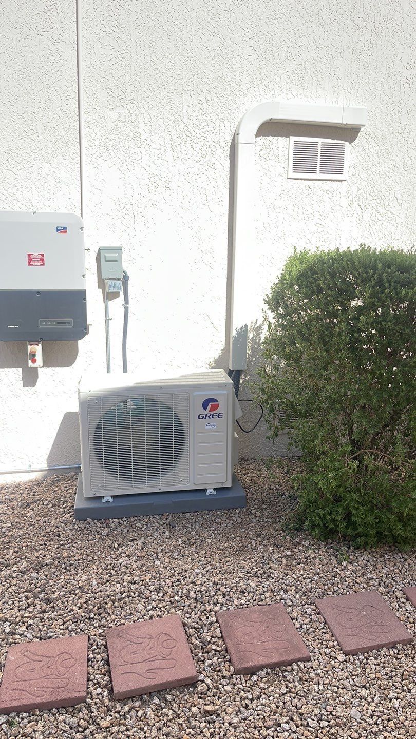 A white air conditioner is sitting outside of a building next to a bush.