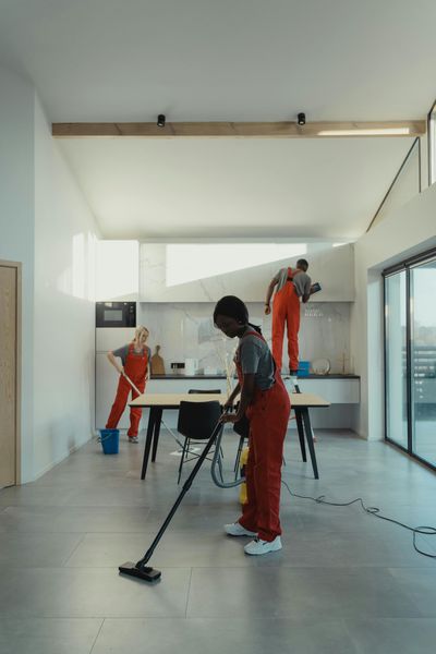 Cleaning crew in red overalls tidying a modern, bright kitchen. One vacuums, another mops, and the third cleans the wall.