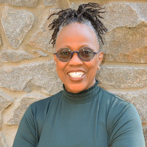 A woman wearing sunglasses and a green shirt is smiling in front of a stone wall.